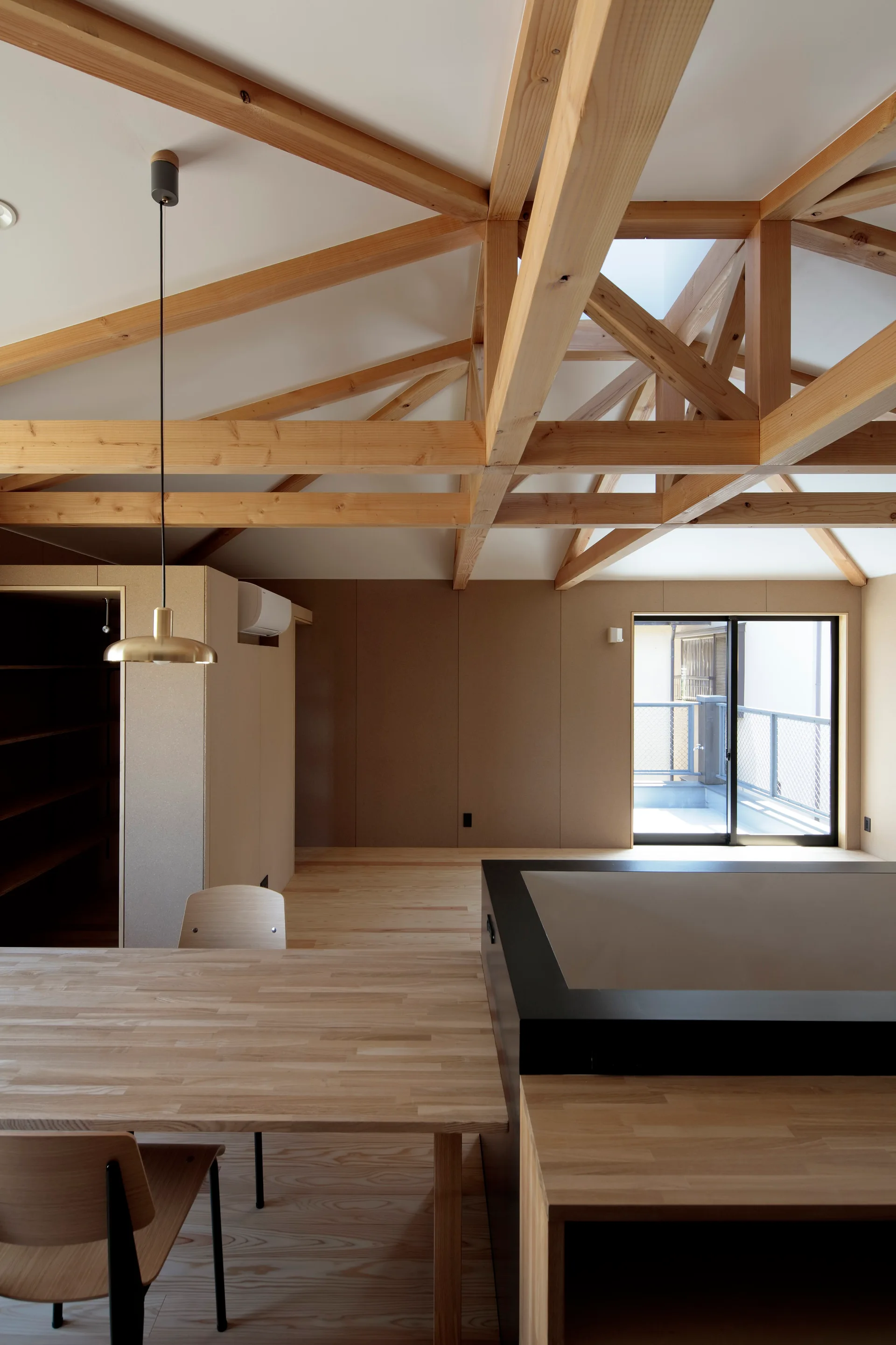 A minimalist dining room with a light wood table, a gold pendant light, and a complex wood truss ceiling under a skylight.