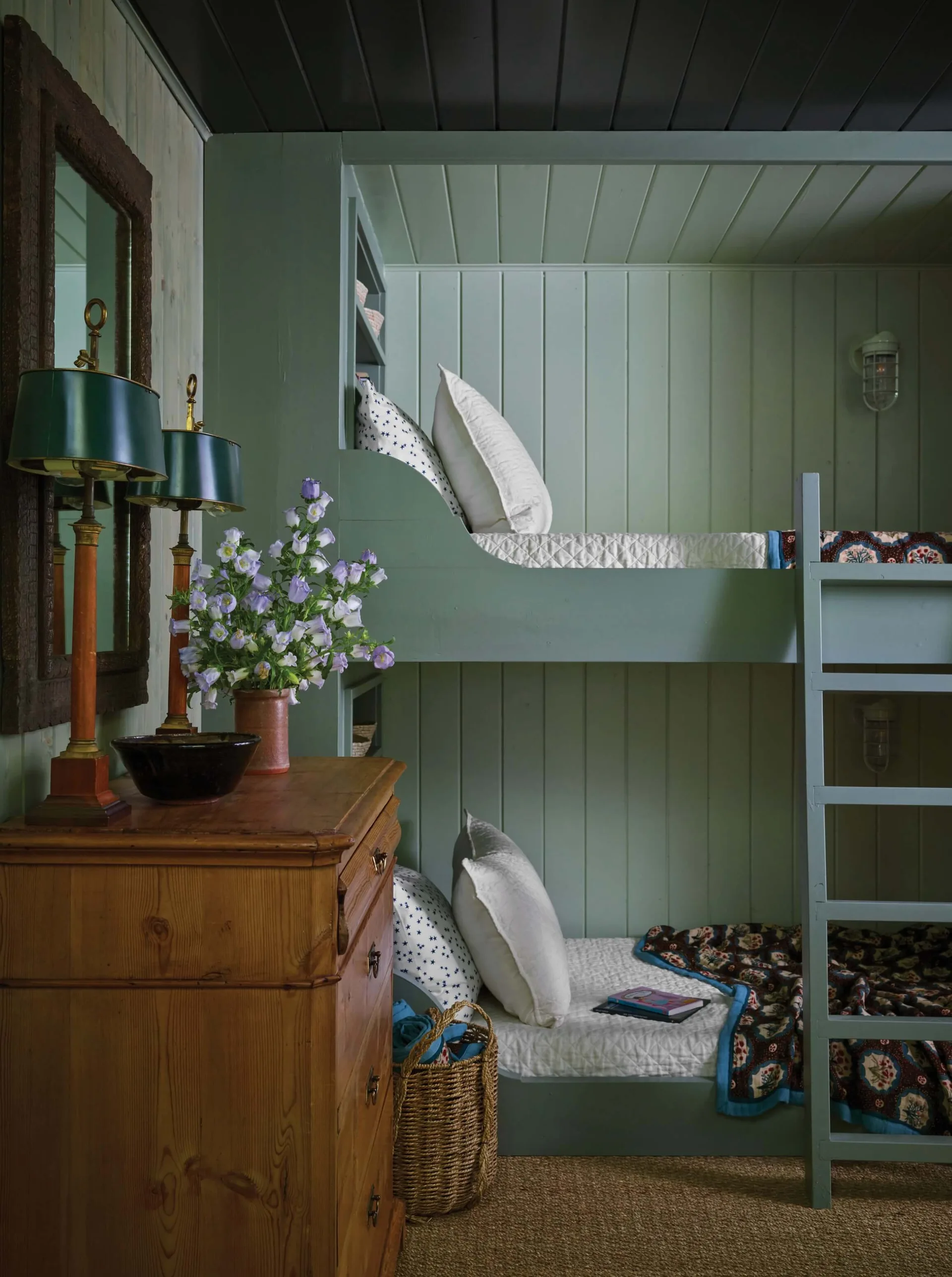 Sage green built-in bunk beds with matching paneled walls stand next to a warm wooden dresser topped with green lamps and purple flowers.