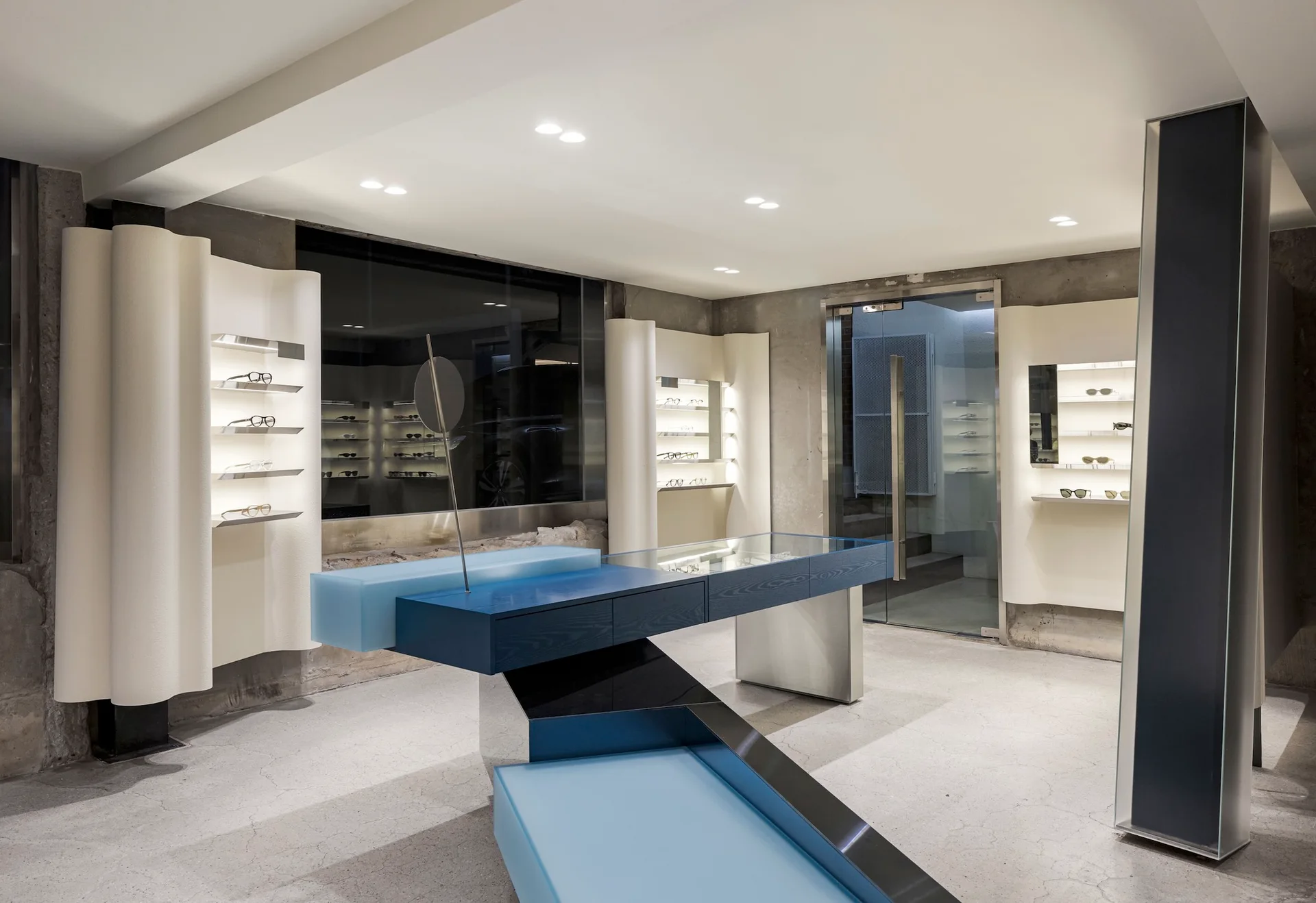 A modern eyewear showroom interior features a sculptural blue geometric display counter, curved white wall shelving units, and raw concrete walls.