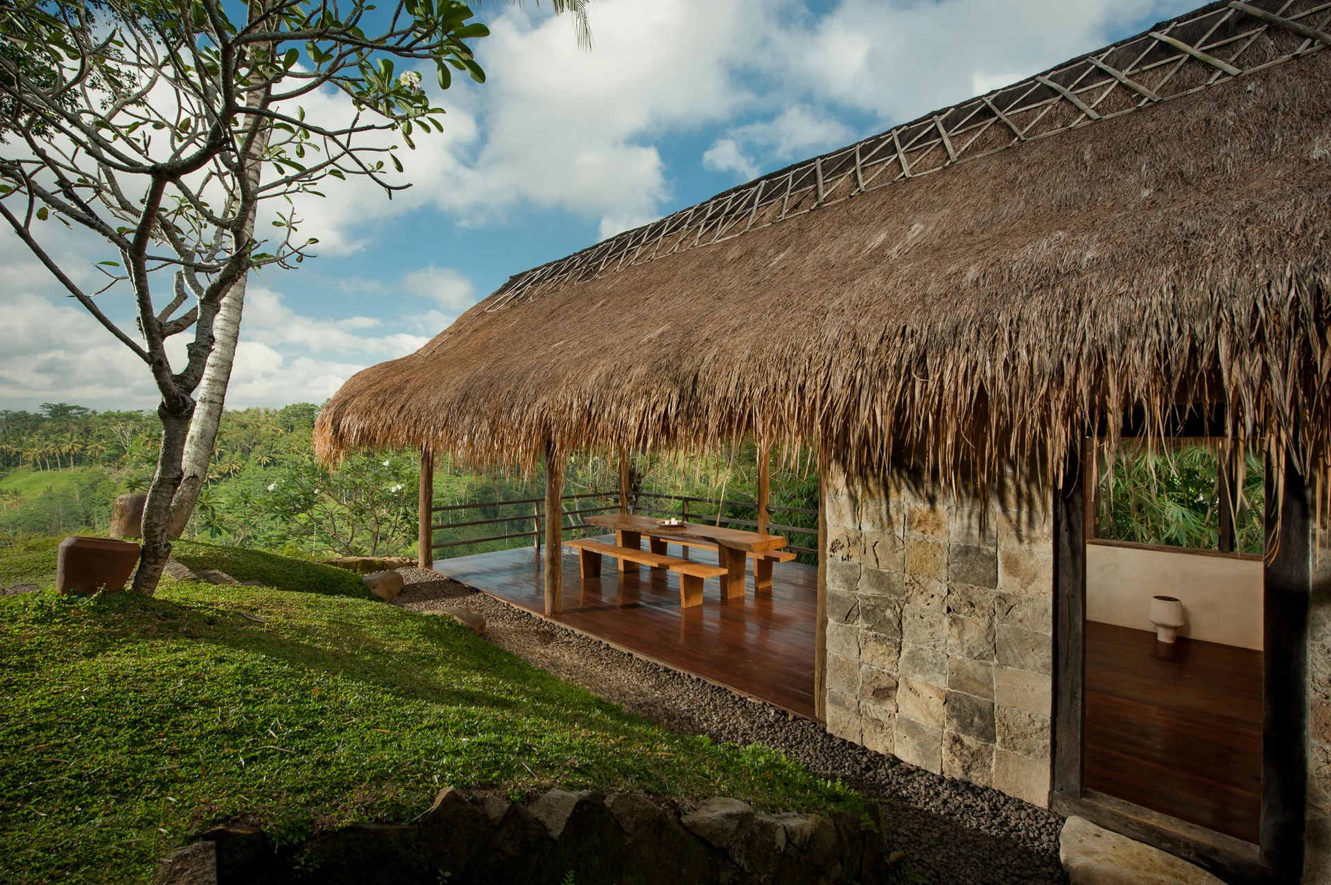 A rustic outdoor dining pavilion features a thatched roof, a stone-block wall, a dark wood deck, and a wooden table with matching benches.