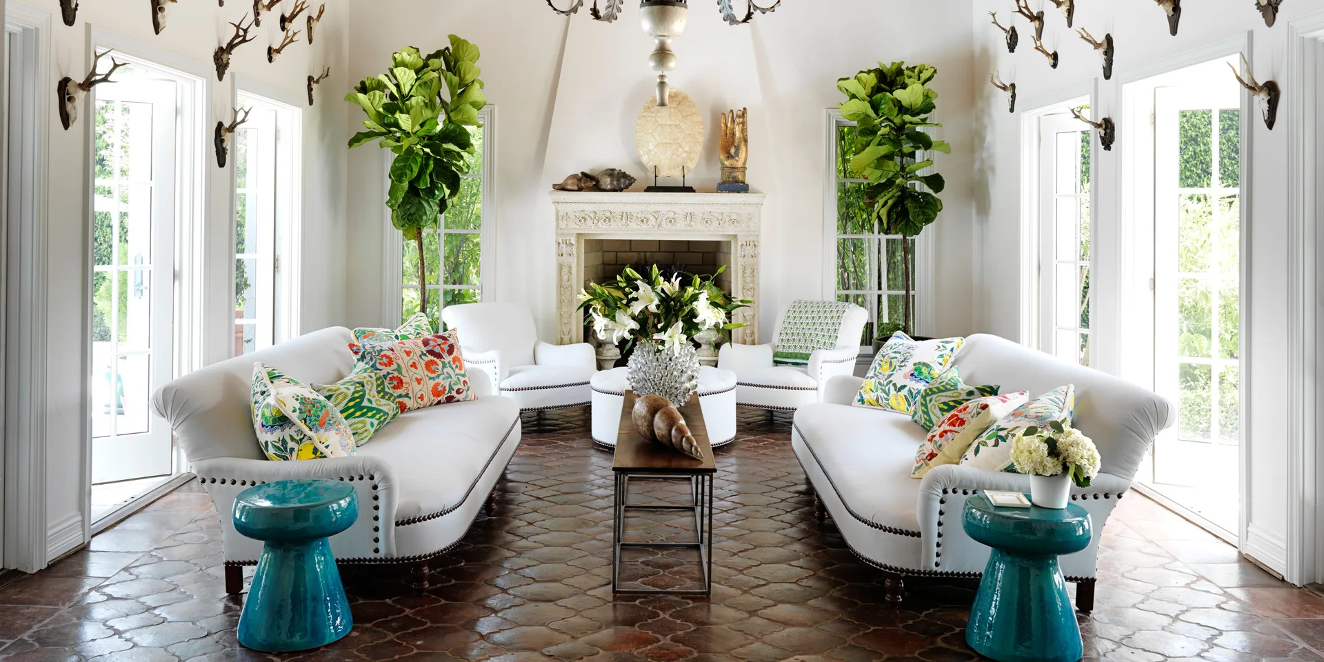 Symmetrical living room featuring white sofas, vibrant teal side tables, and a carved stone fireplace set on patterned terracotta tile flooring.