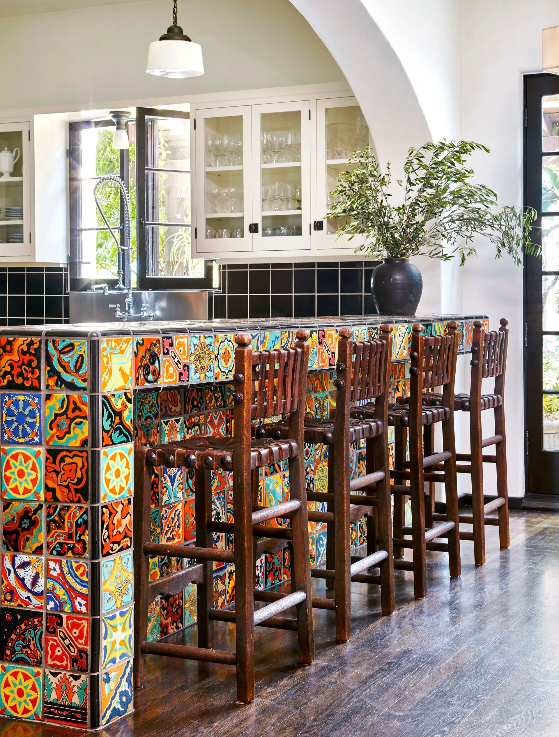 A kitchen island wrapped in multicolored patterned ceramic tiles stands next to dark wooden bar stools on a polished dark wood floor.