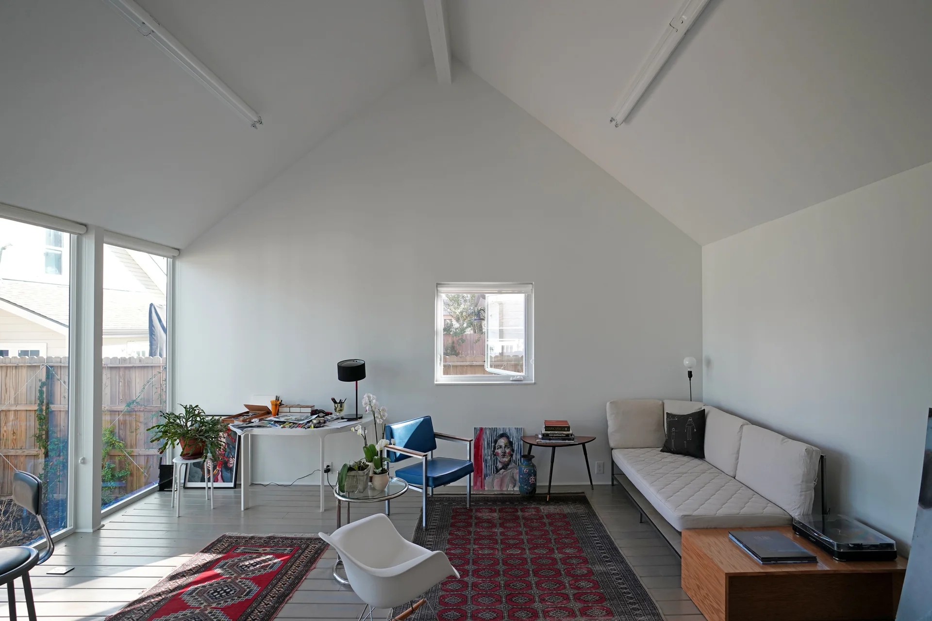 A bright living room with a vaulted ceiling features a white sofa, a blue armchair, a desk, and red patterned rugs on grey flooring.