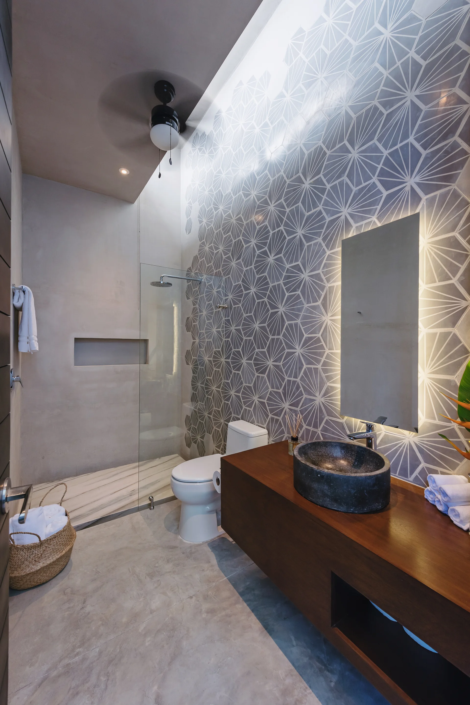 A modern bathroom featuring a dark stone vessel sink on a wood floating vanity beside a glass shower and a grey geometric tiled accent wall.