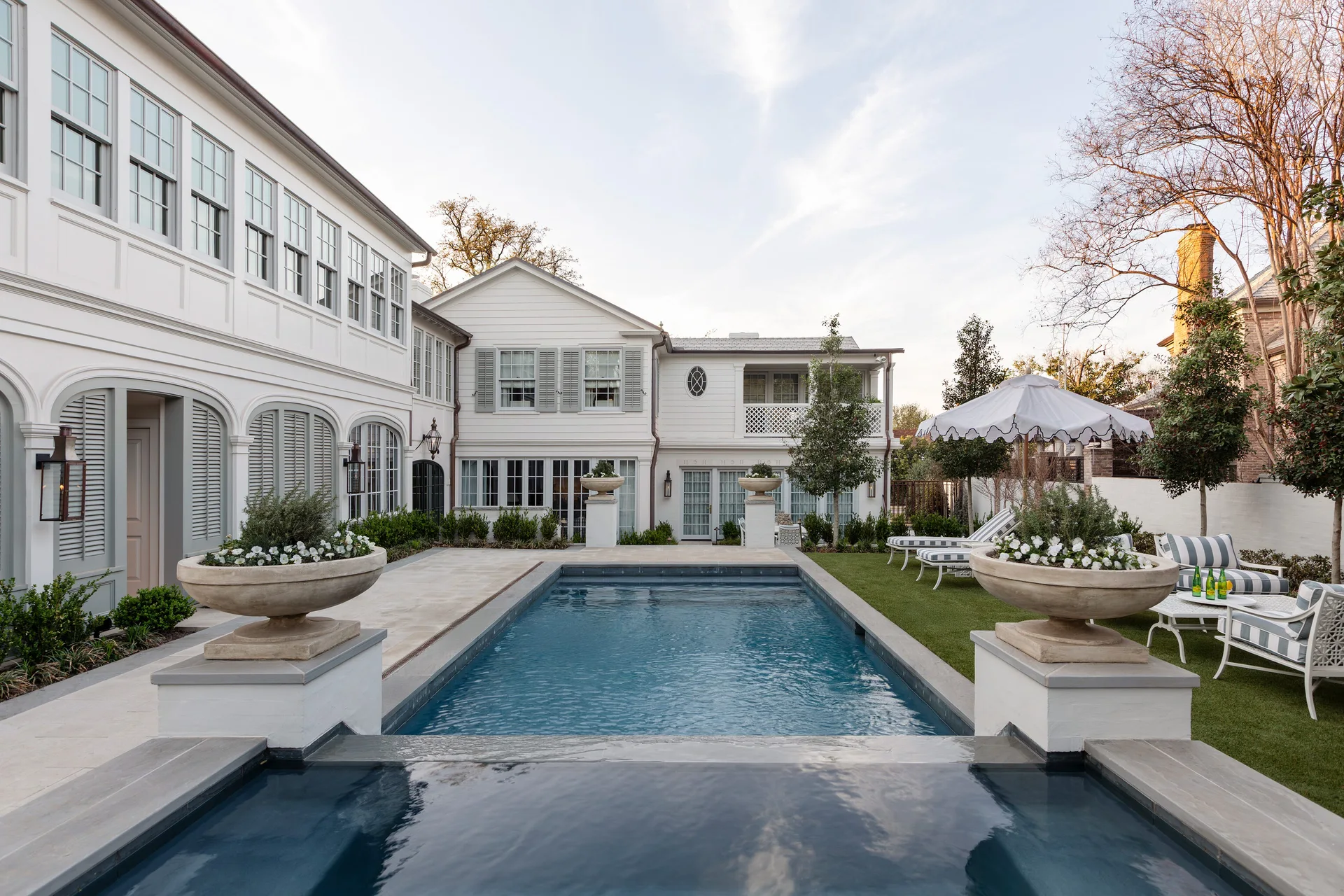 A rectangular swimming pool is flanked by stone urn planters on pedestals in a landscaped backyard next to a white shuttered house.