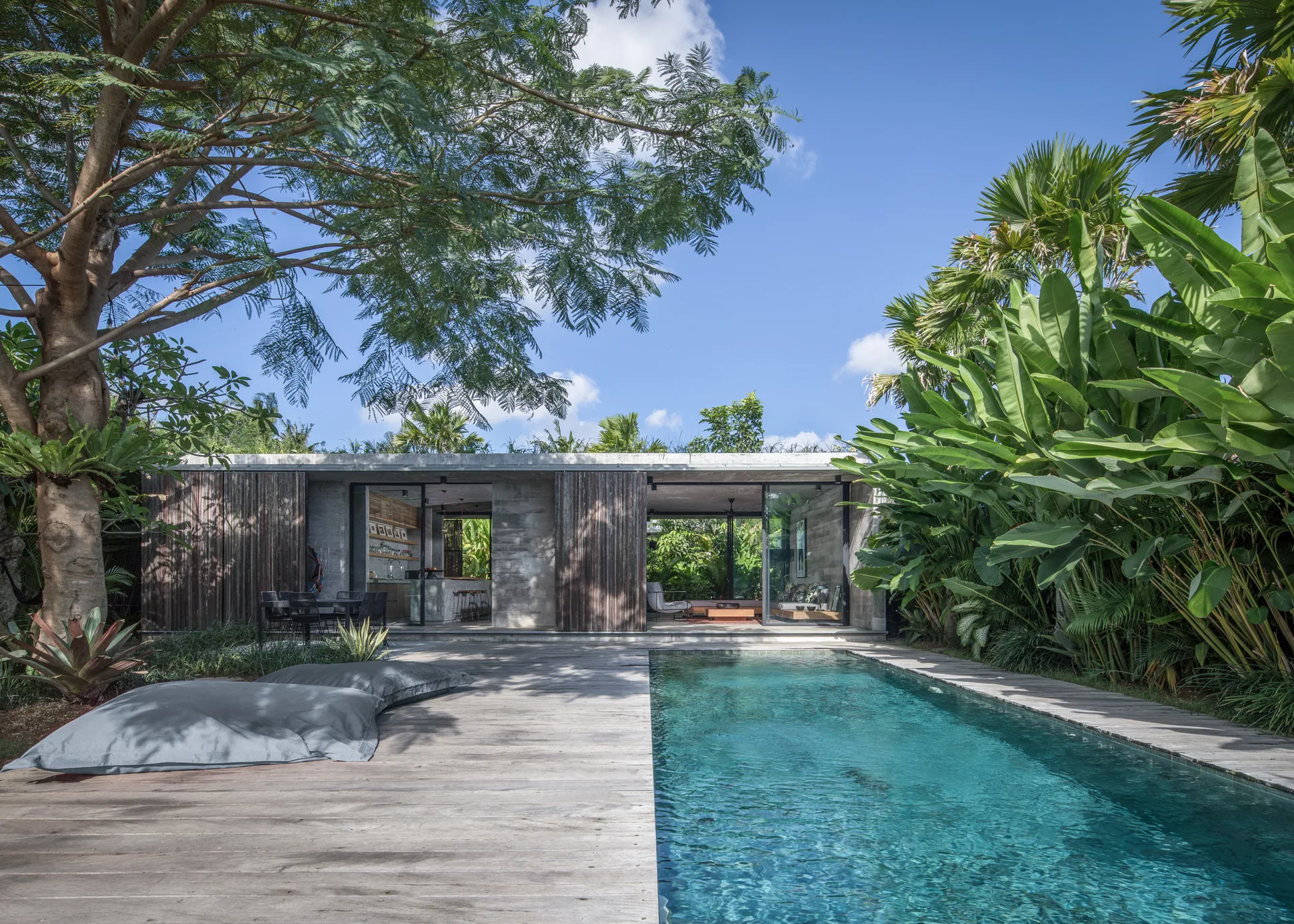 A modern concrete pavilion features a lap pool, a weathered wooden deck with bean bags, and sliding glass walls surrounded by tropical foliage.