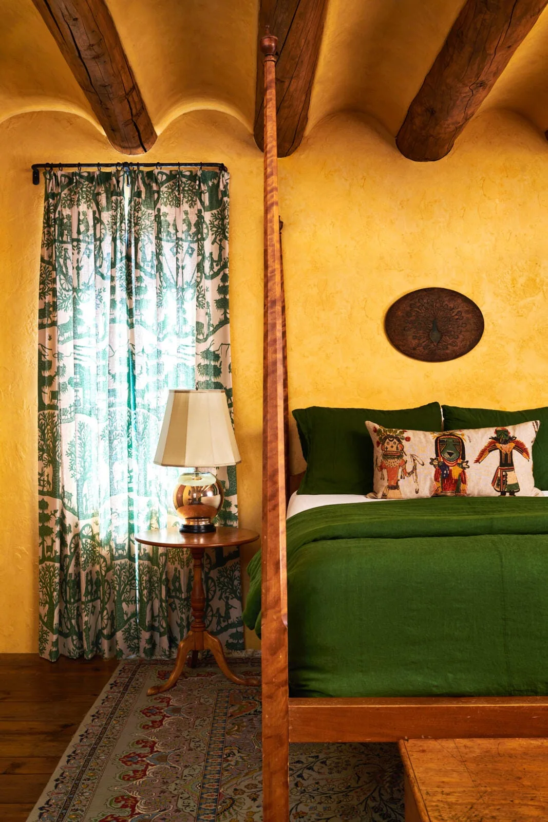 A rustic bedroom features textured yellow plaster walls, exposed wooden ceiling beams, and a wood four-poster bed with green linens.