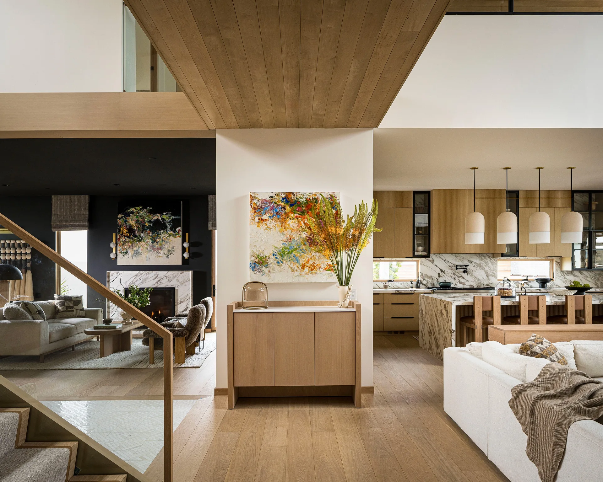 An open-concept home interior featuring a wood console table beneath abstract art, a fireplace lounge to the left, and a marble kitchen to the right.