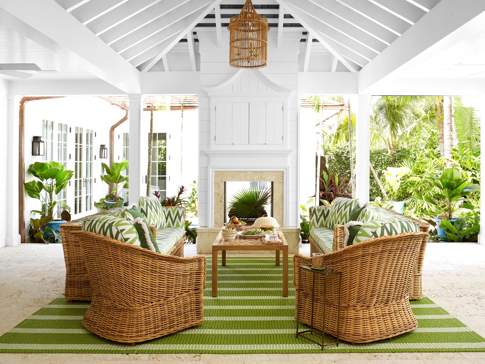 A covered patio features wicker sofas with green and white patterned cushions, a central stone fireplace, and a green striped rug.