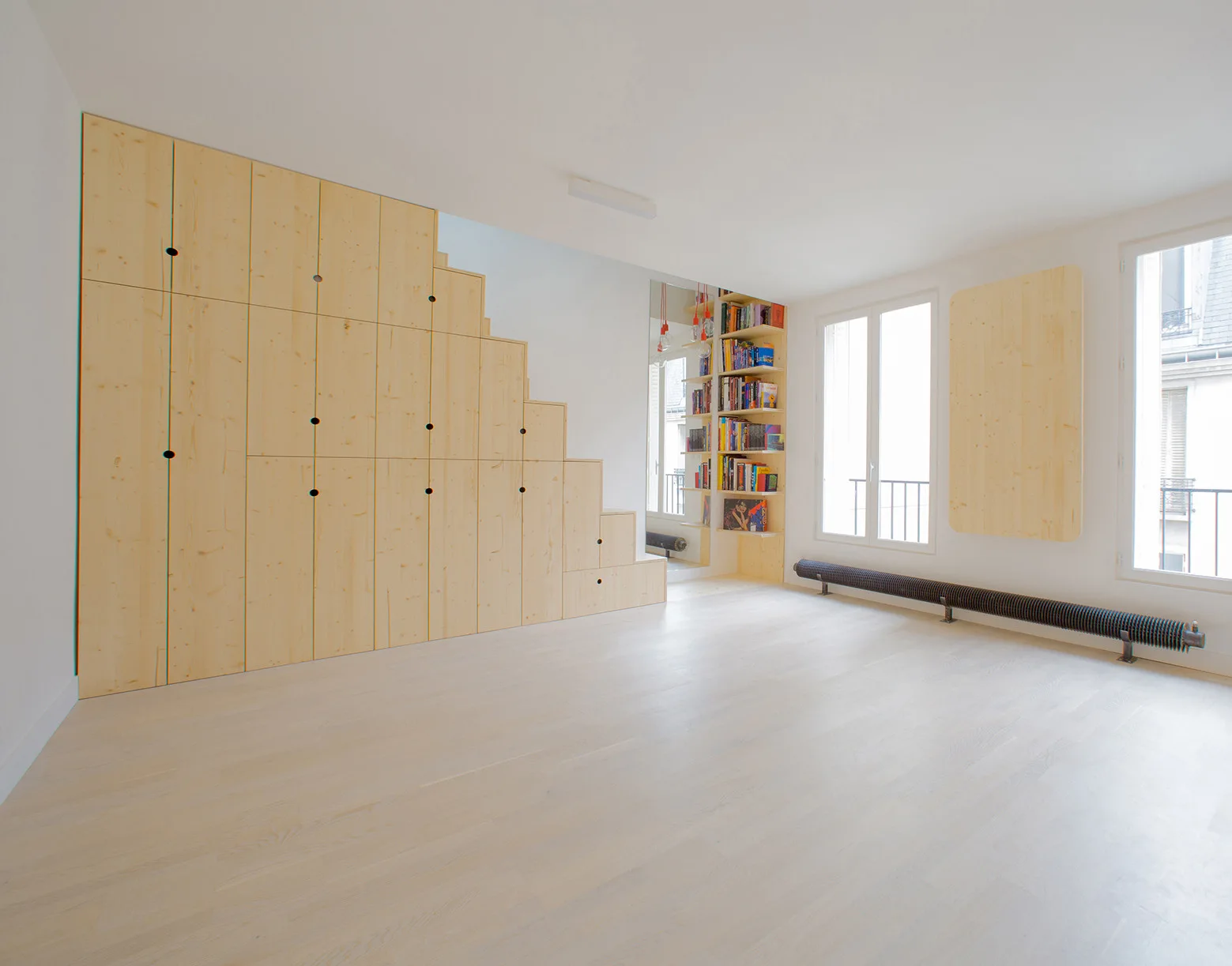 A bright room features light parquet flooring, a large stepped wooden storage cabinet, and two tall windows next to an industrial radiator.