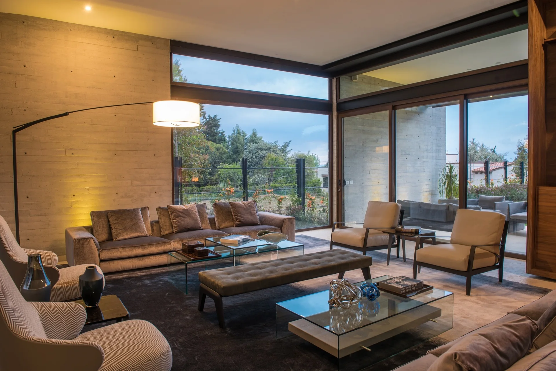 A modern living room features a board-formed concrete wall, a velvet sofa, leather armchairs, glass coffee tables, and large windows.