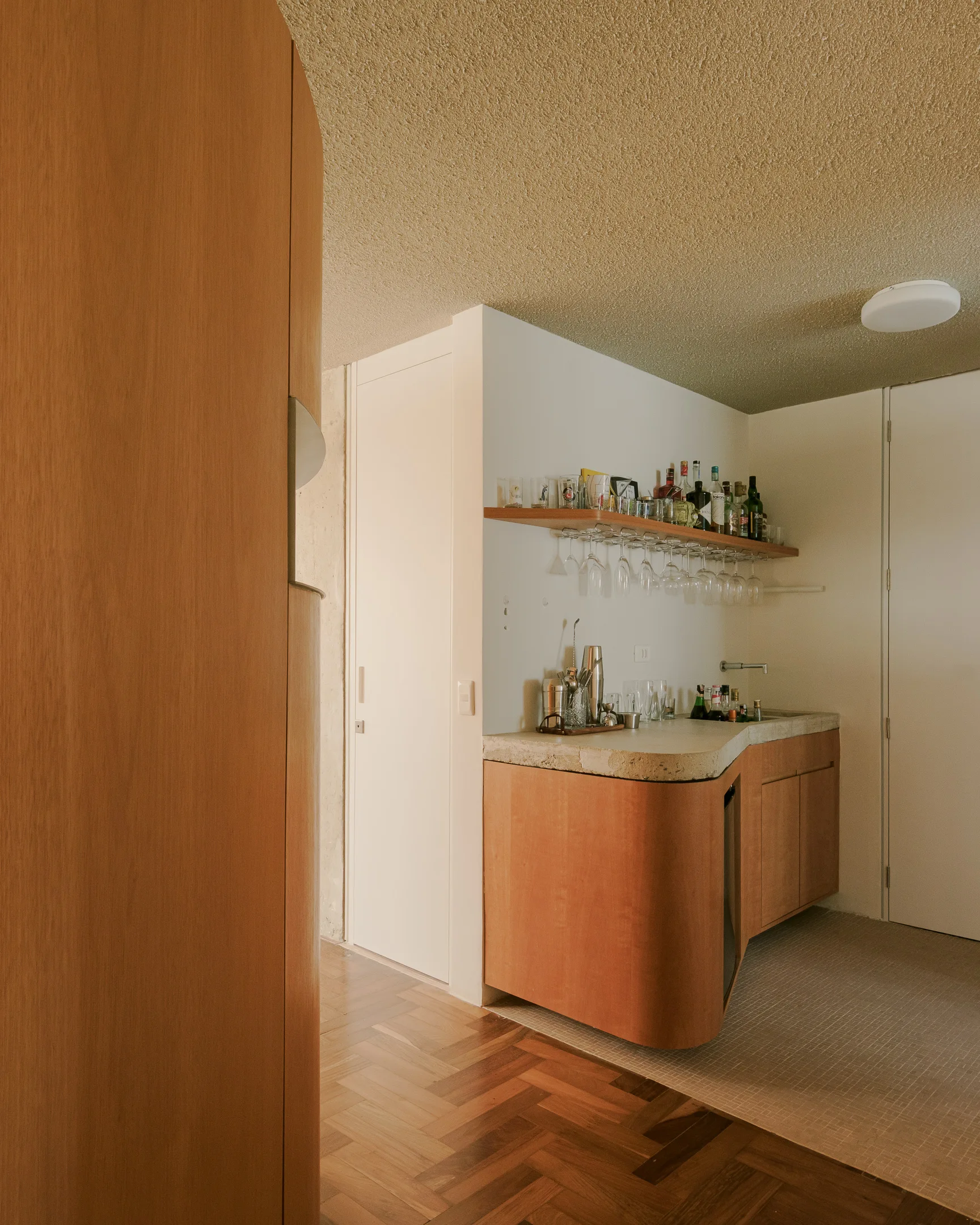 A compact wet bar features a curved wood cabinet with a stone-textured countertop, a floating wooden shelf holding glassware, and herringbone wood flooring.