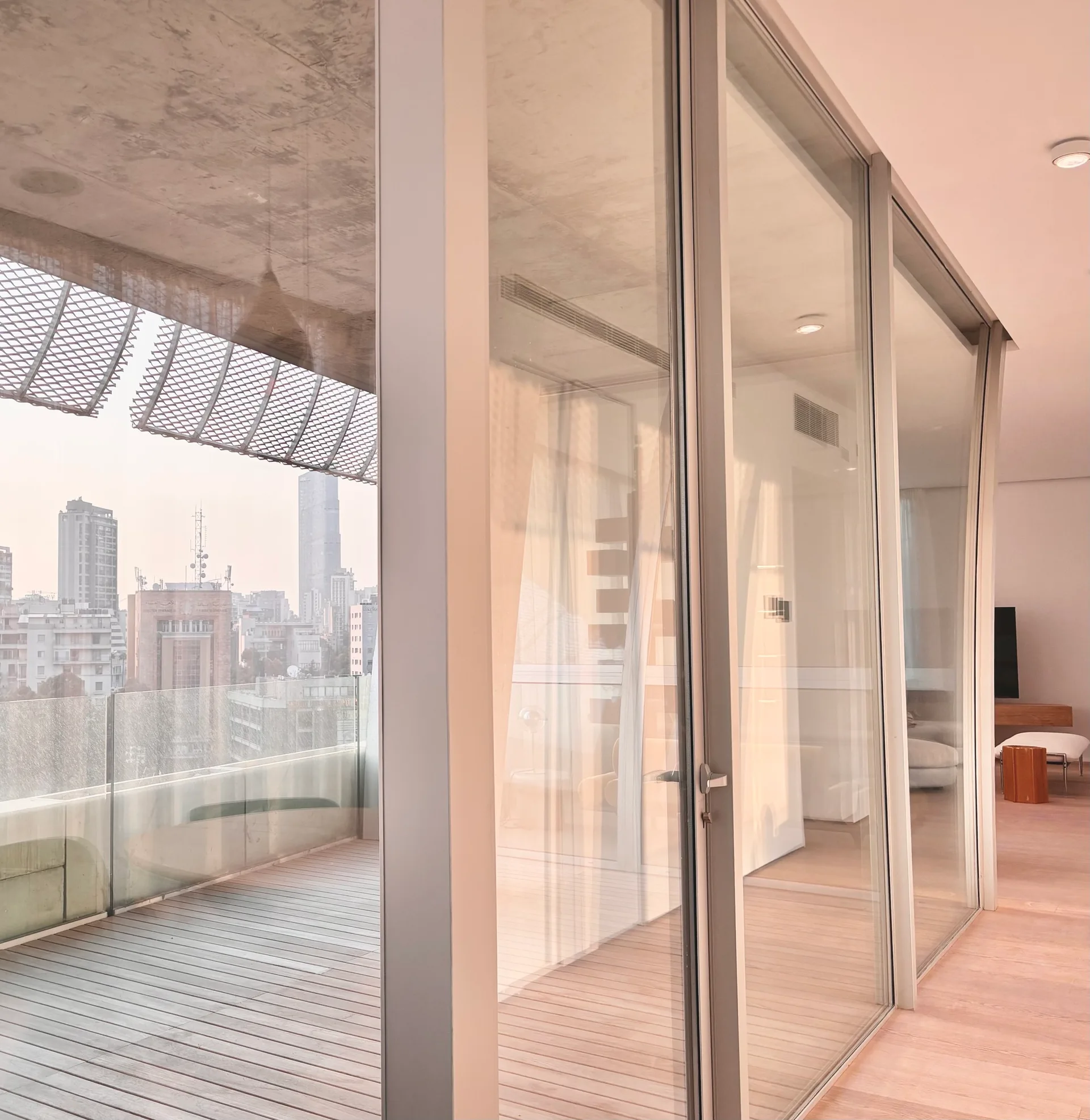 Floor-to-ceiling glass sliding doors separate a minimalist interior room from an outdoor balcony with wood decking and a view of city buildings.