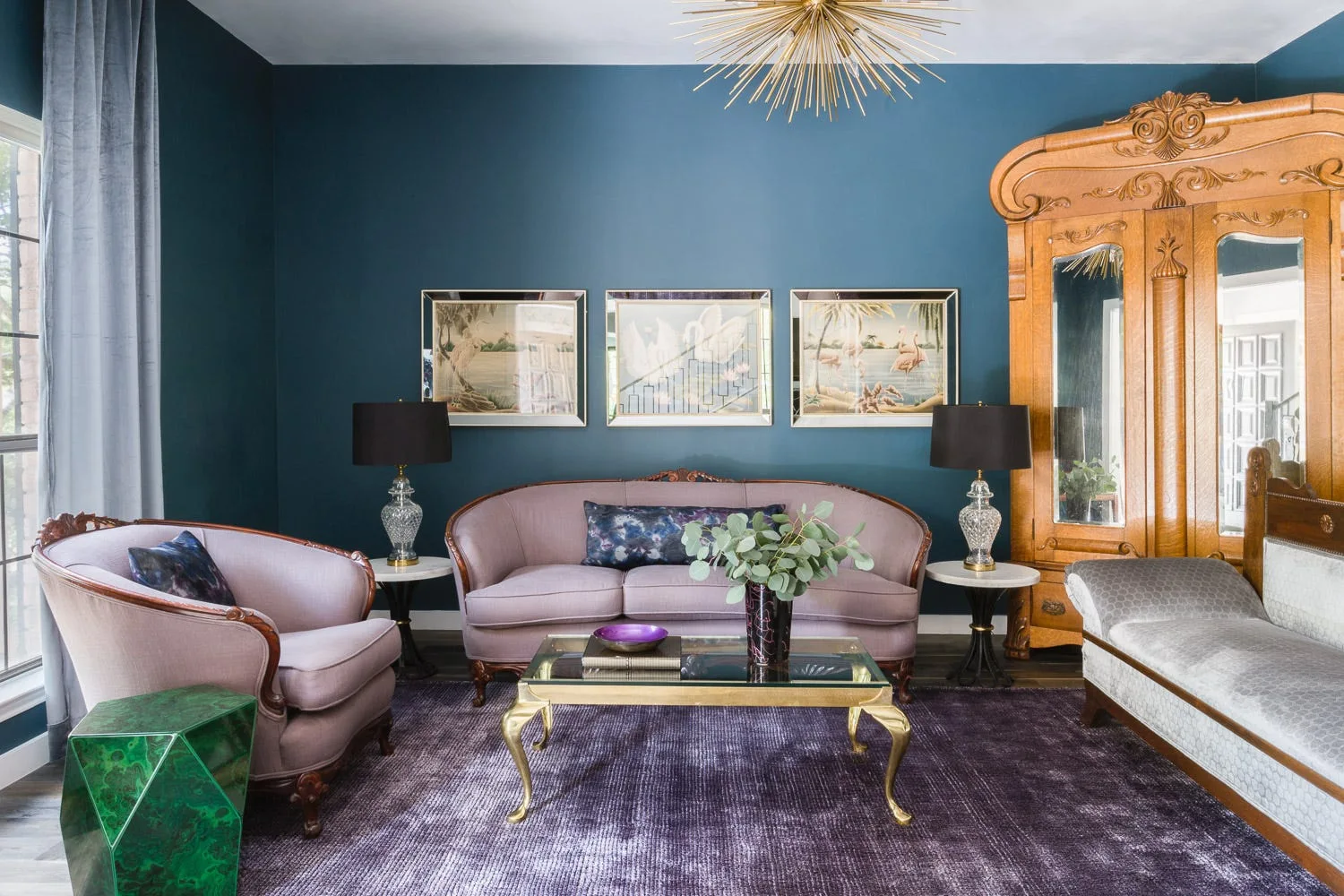 A blue living room features lilac upholstered sofas, a textured purple rug, a large wooden armoire, and a gold starburst chandelier.