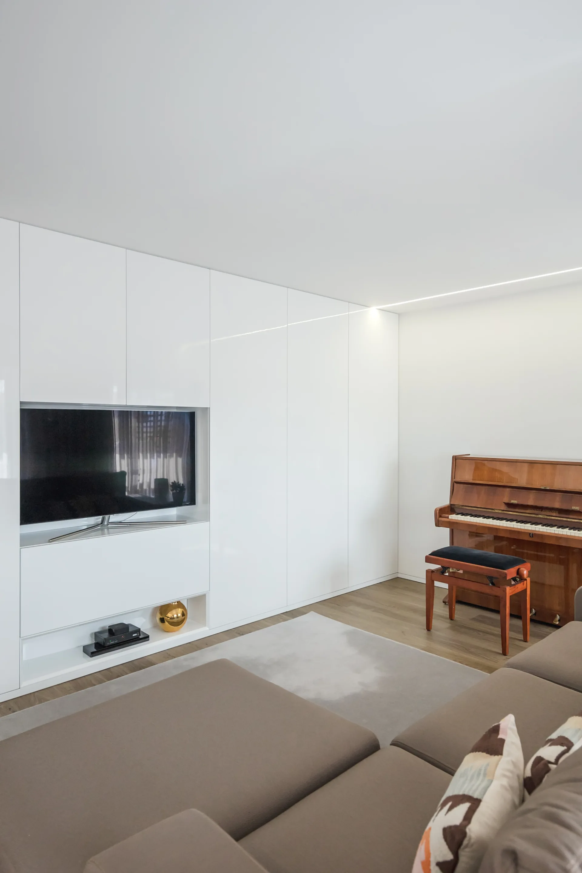 A modern living room features a large white built-in media cabinet with an integrated TV next to a wood upright piano and a brown sectional sofa.