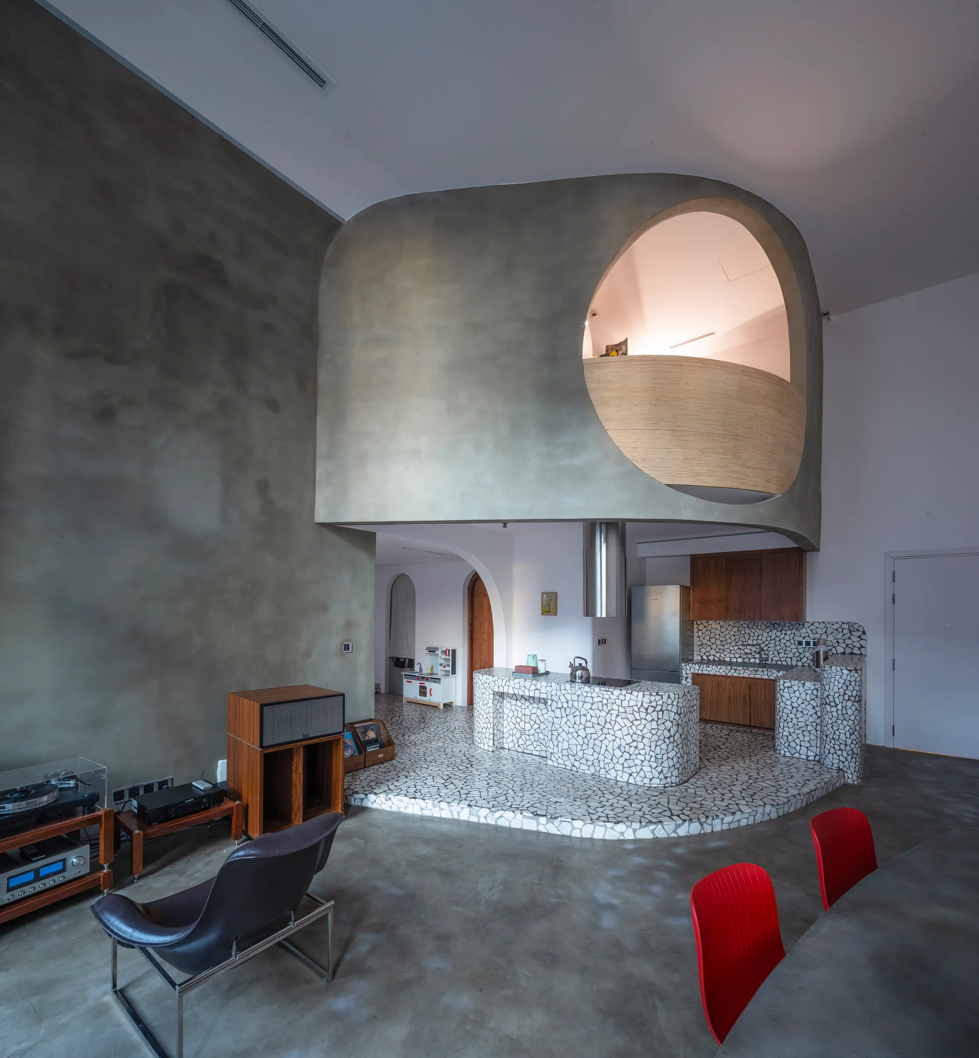 An open-plan living space features a curved concrete mezzanine loft above a kitchen finished with white stone mosaic tiles.
