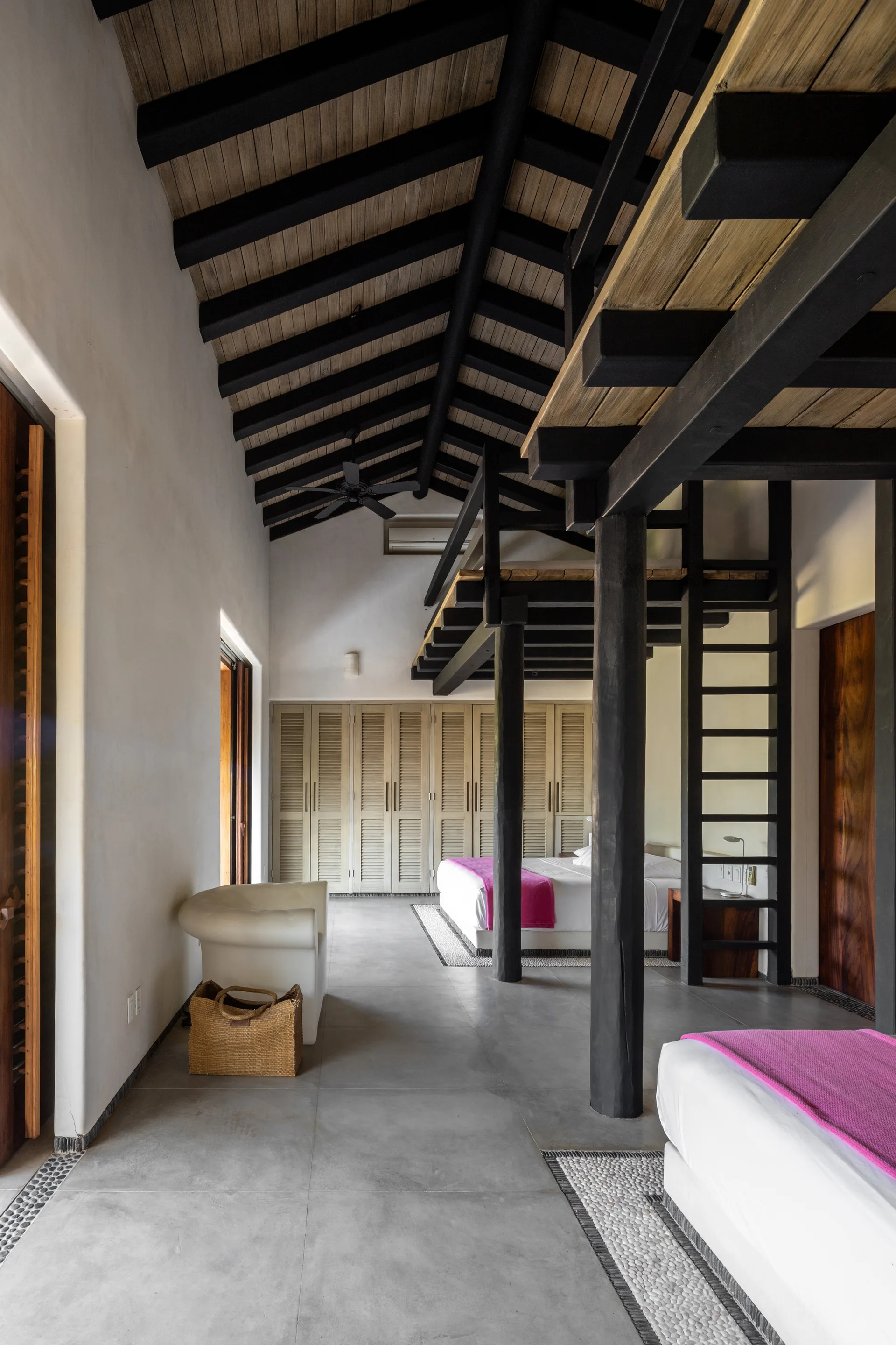 A spacious loft bedroom with a high ceiling, dark wood rafters, two white beds with hot pink throws, and a black ladder.