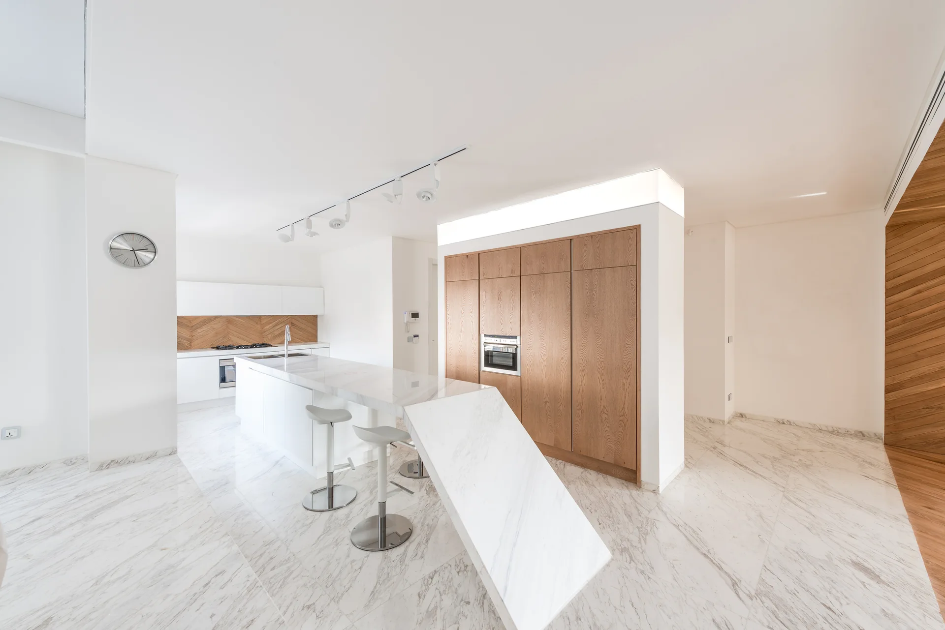 A modern minimalist kitchen featuring a white marble waterfall island with an angled edge, silver bar stools, and integrated wood cabinets.