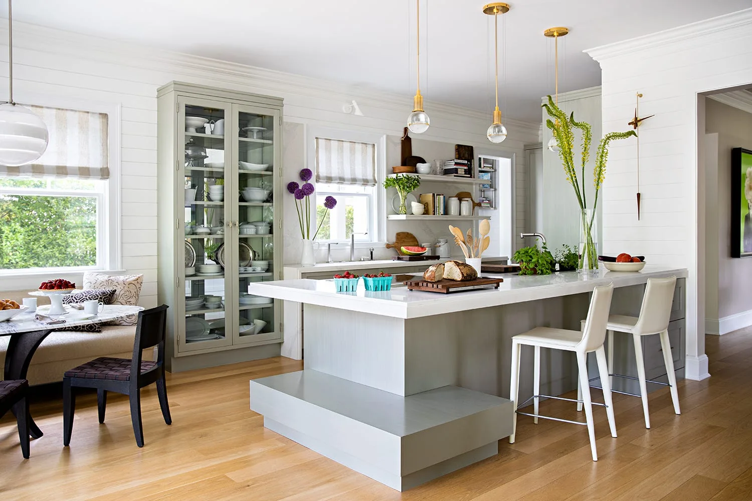 A modern farmhouse kitchen featuring white shiplap walls, a central gray island with white bar stools, and a tall sage green glass-front cabinet.