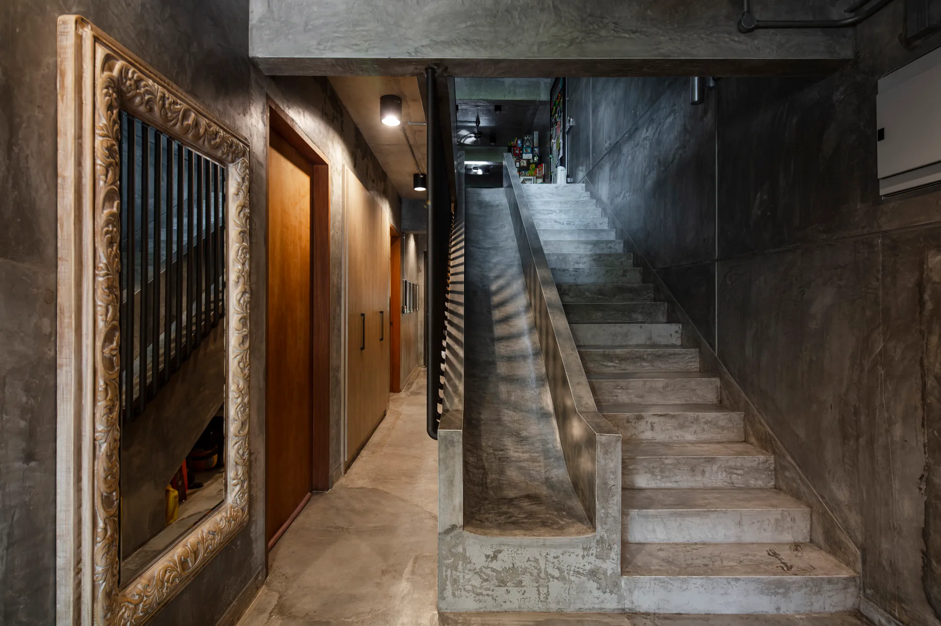 A concrete staircase with an integrated slide stands next to a hallway with wooden doors and a large ornate wood-framed mirror on a textured concrete wall.