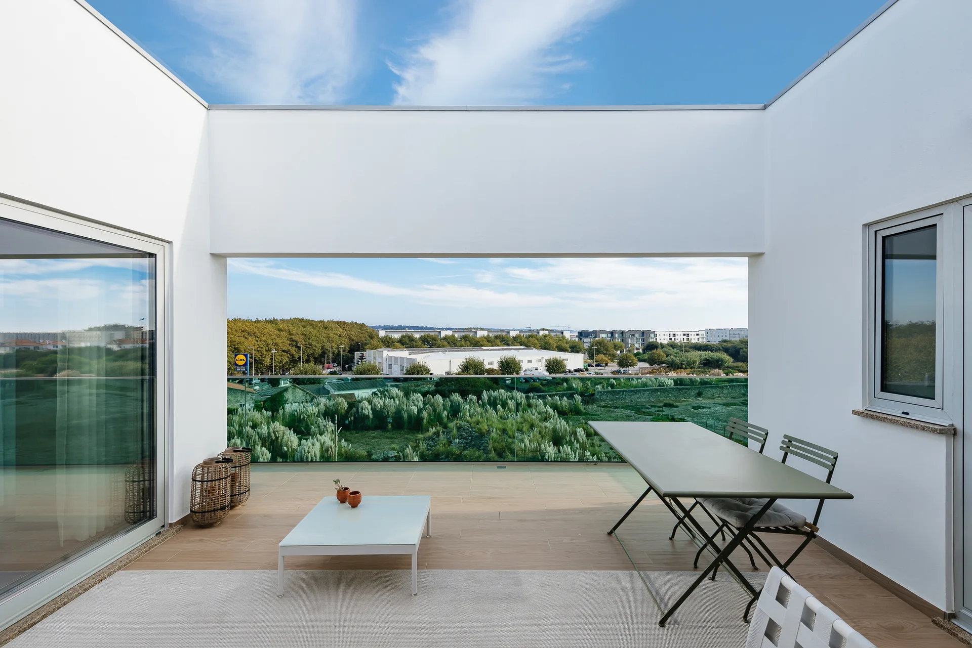 A modern white terrace features a glass railing, a green metal dining table with chairs, a low white coffee table, and wood-look plank flooring.