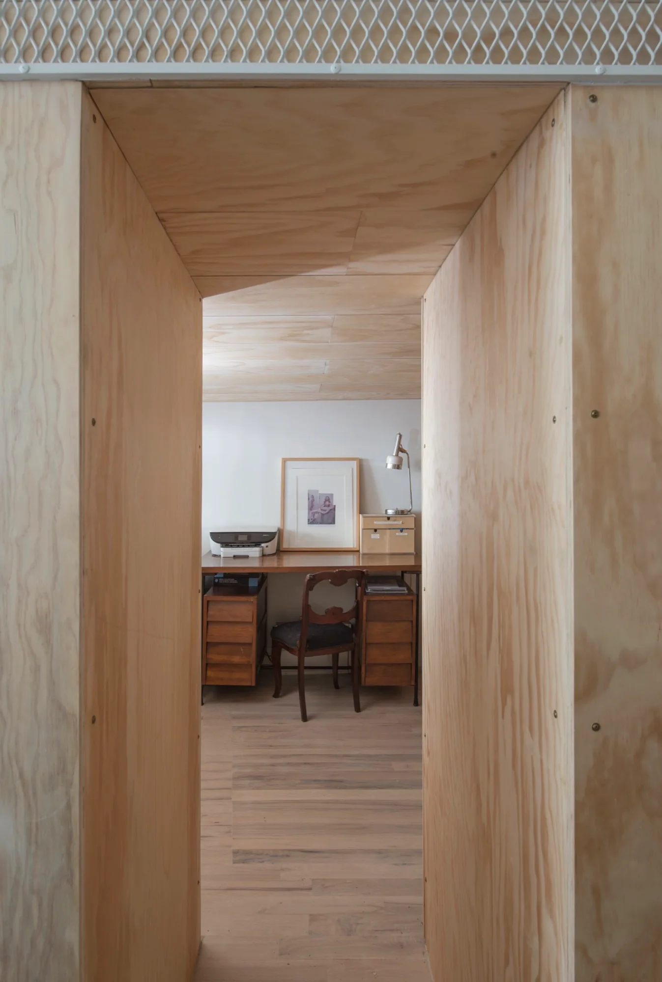 A view through a deep plywood-clad doorway reveals a quiet home office with a wooden desk, antique chair, and simple desk accessories.
