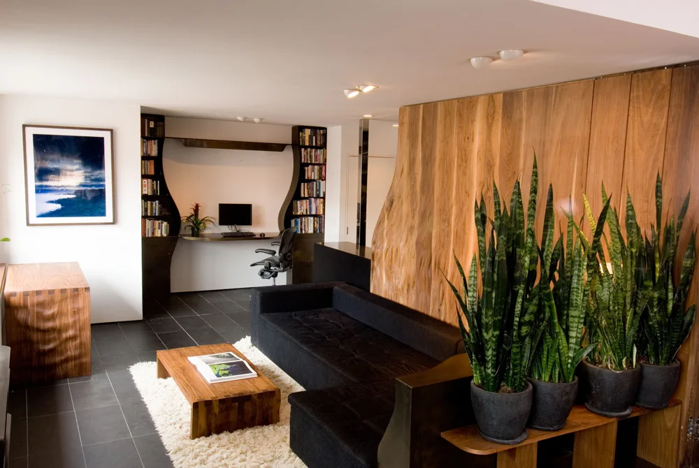 A contemporary living room featuring a black sectional sofa, a live-edge wood feature wall, potted snake plants, and a built-in office desk.