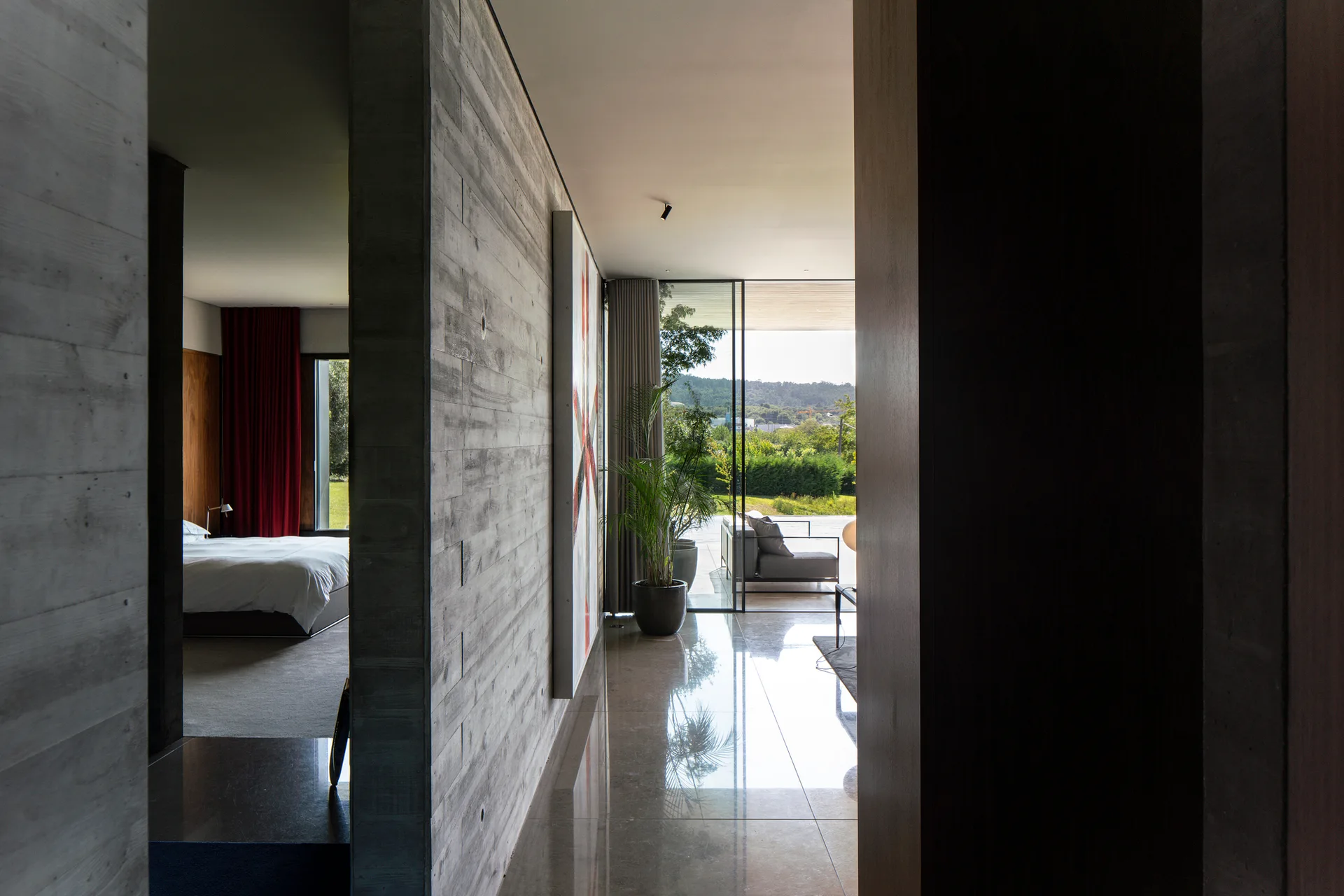 A modern hallway with board-formed concrete walls and polished stone floors leading to a glass-walled patio and a bedroom with red curtains.