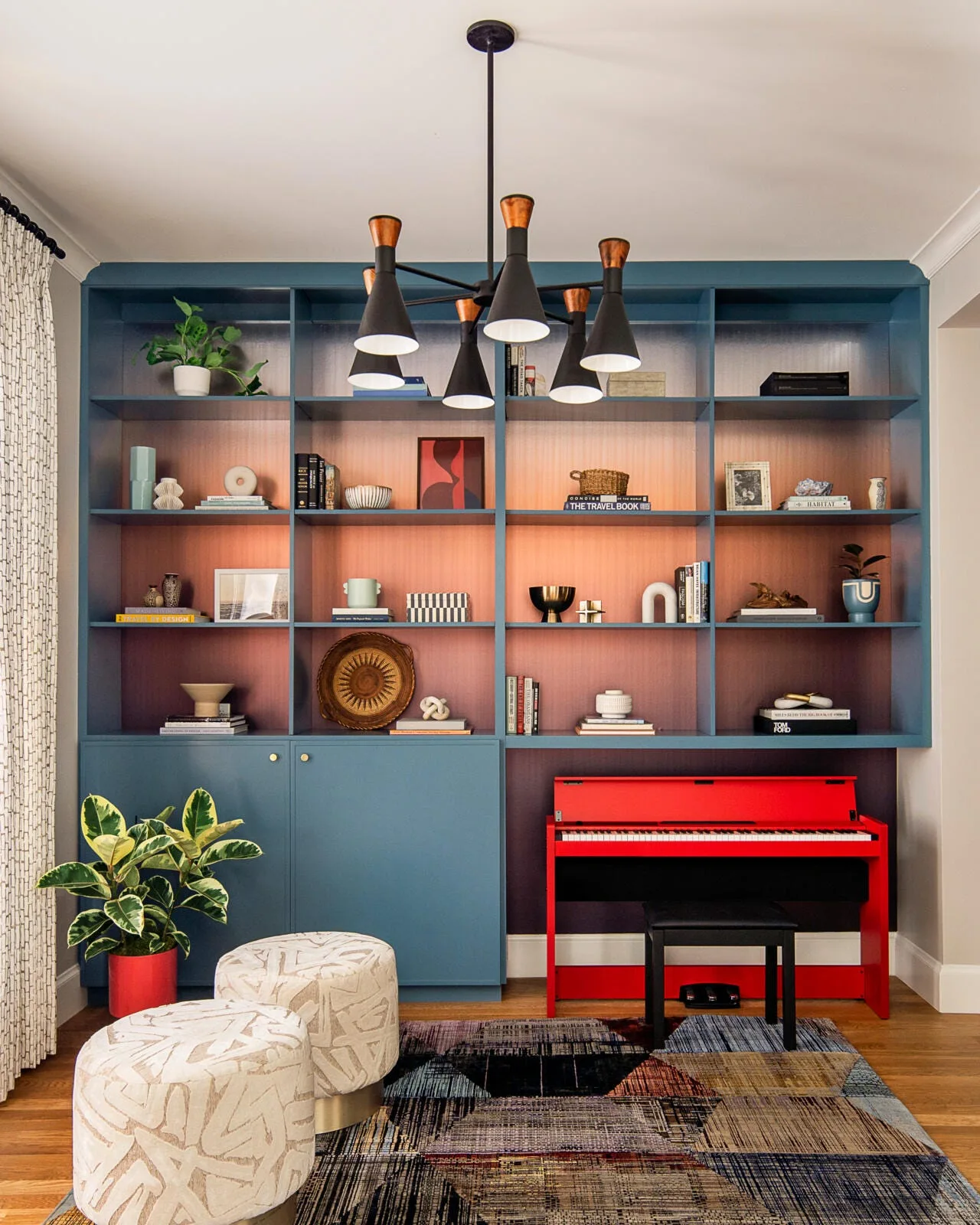 A large blue custom built-in bookcase features an ombré sunset-toned back panel, curated ceramic decor, and an integrated bright red piano.