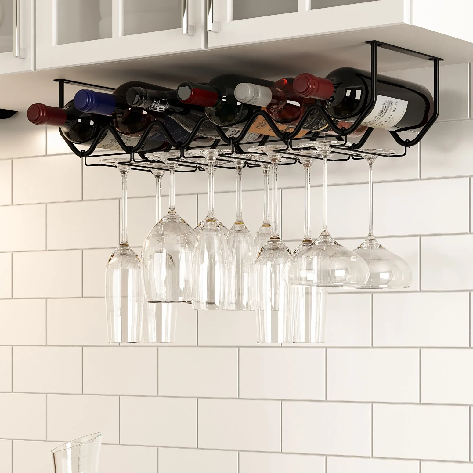 A black metal rack mounted beneath white kitchen cabinets holds wine bottles and hanging stemware against a white subway tile backsplash.