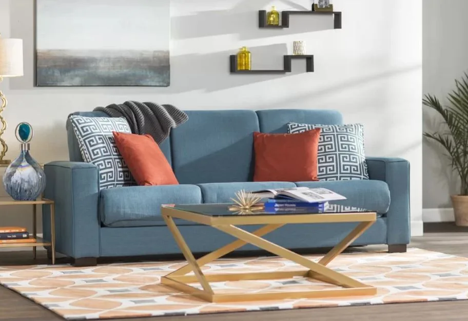 A contemporary blue fabric sofa sits on a patterned rug behind a gold geometric coffee table with a glass top.