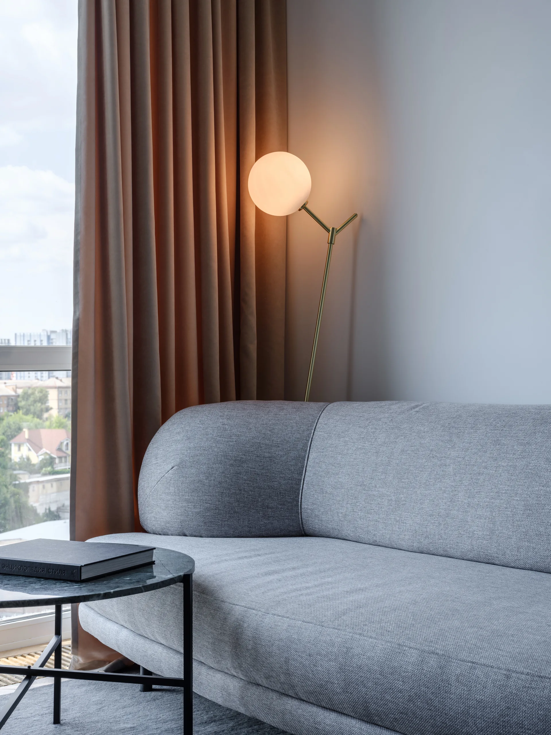 A grey upholstered sofa sits next to a black stone side table and a brass floor lamp with a glowing globe shade in front of brown curtains.