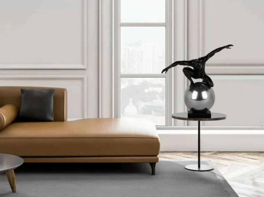 A tan leather chaise lounge sits next to a black pedestal table holding a chrome sphere sculpture in front of a window and paneled wall.