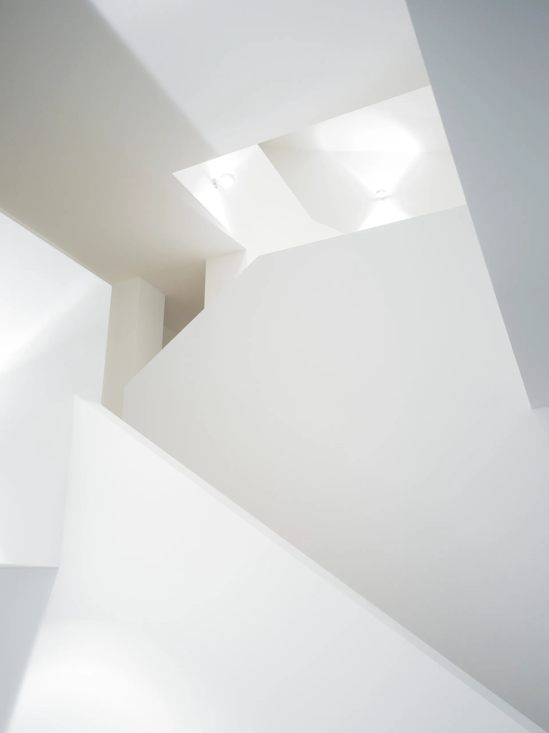 Looking up a minimalist, all-white staircase with sharp geometric walls and small recessed spotlights in the ceiling.
