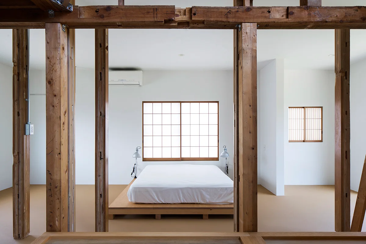 A minimalist bedroom features a low wooden platform bed positioned in front of a shoji grid window, viewed through exposed timber structural posts.