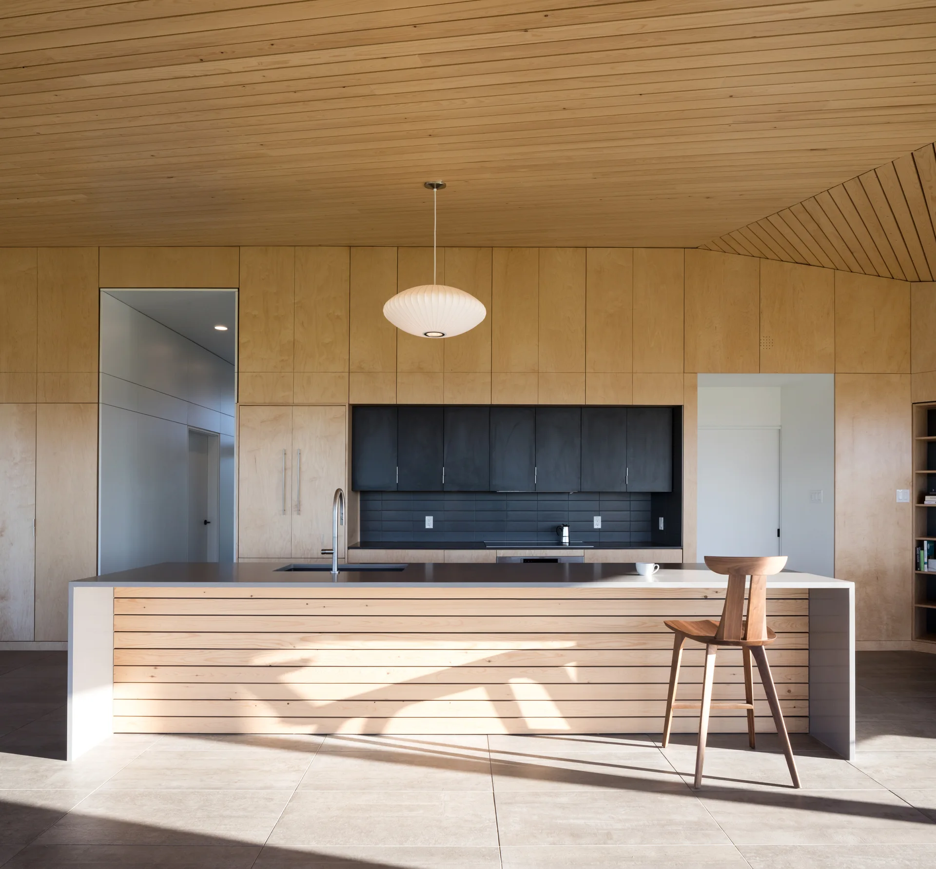 A minimalist kitchen features light wood paneling on the walls and ceiling, a slatted wood island with a gray countertop, and dark matte upper cabinets.