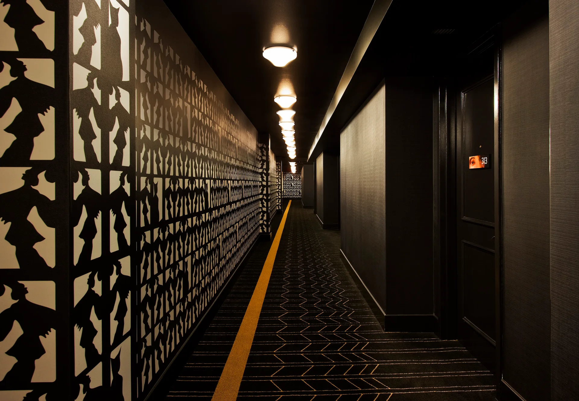 A long dark hallway features black and white silhouette wallpaper on one wall and a patterned black carpet with a single yellow stripe.