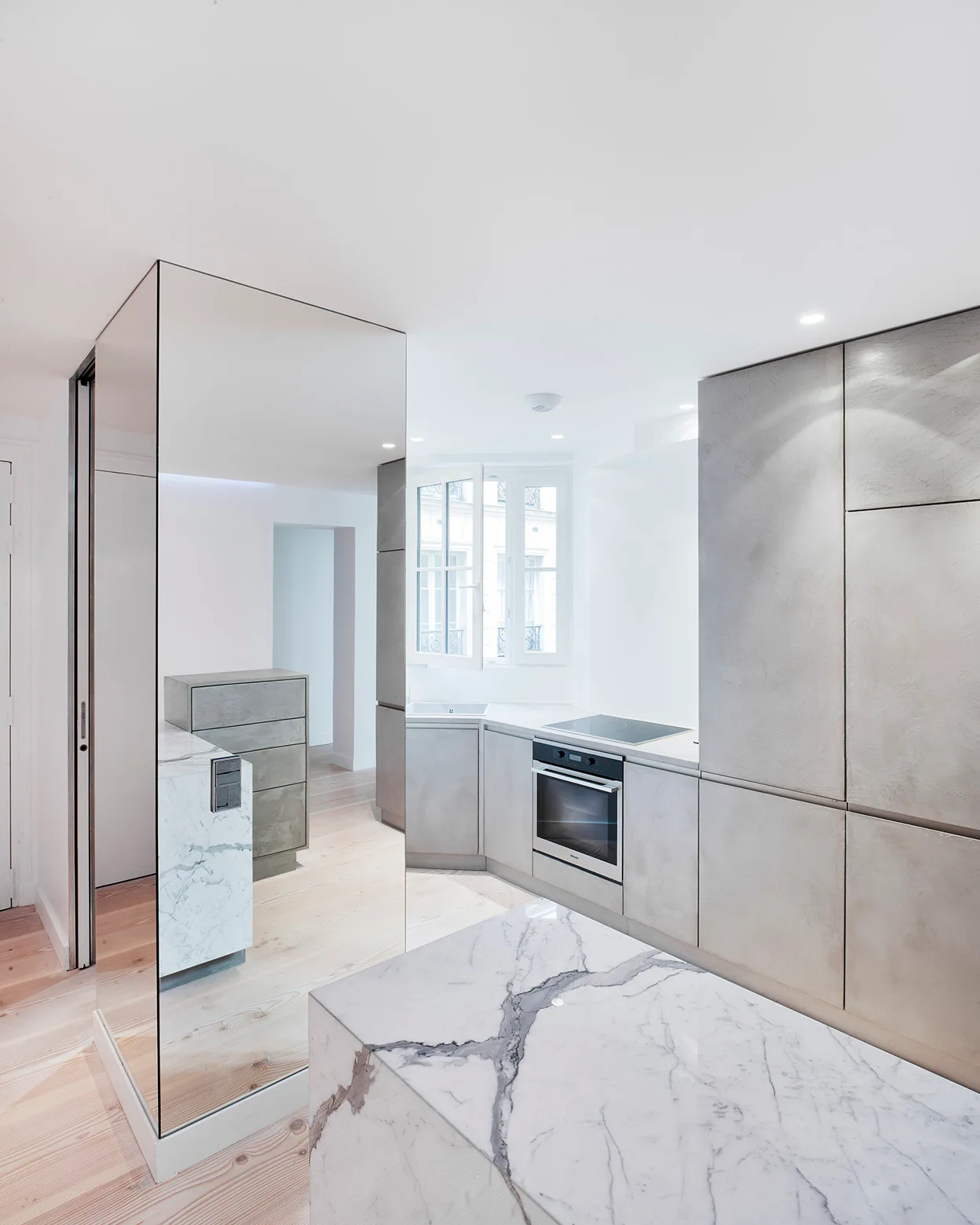 A minimalist kitchen featuring grey integrated cabinetry, a white marble island, light wood plank floors, and a floor-to-ceiling mirrored panel.