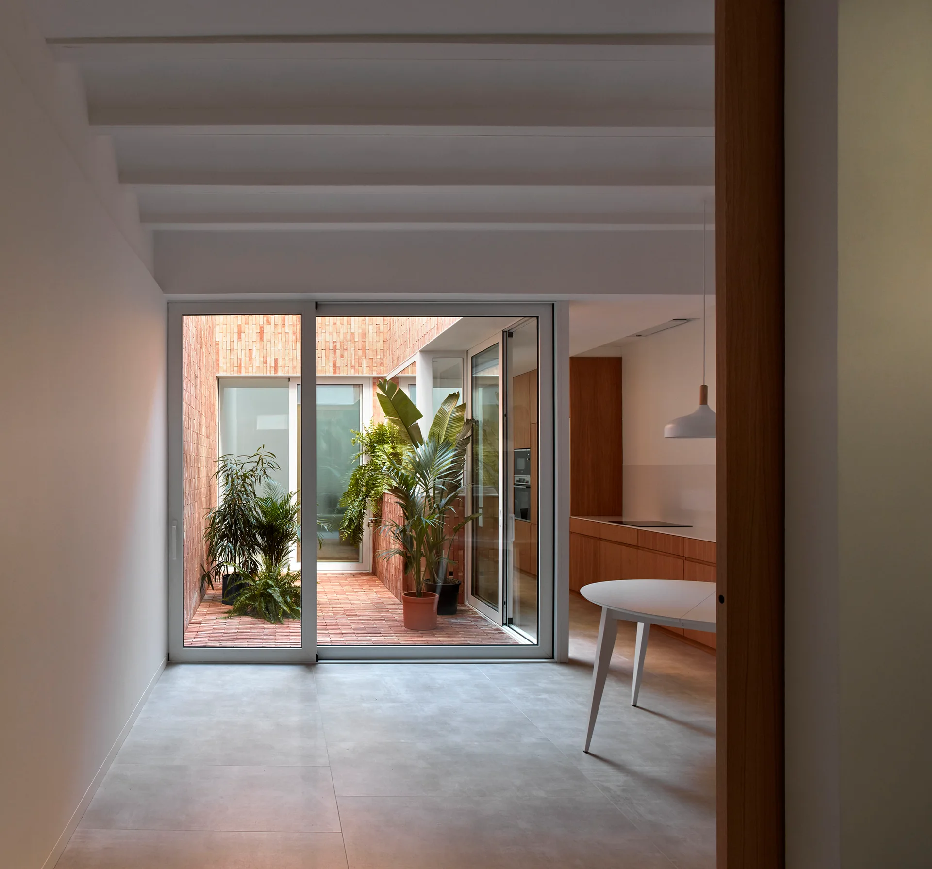 A modern home interior with grey tile flooring features sliding glass doors opening to a brick-paved courtyard filled with potted green plants.
