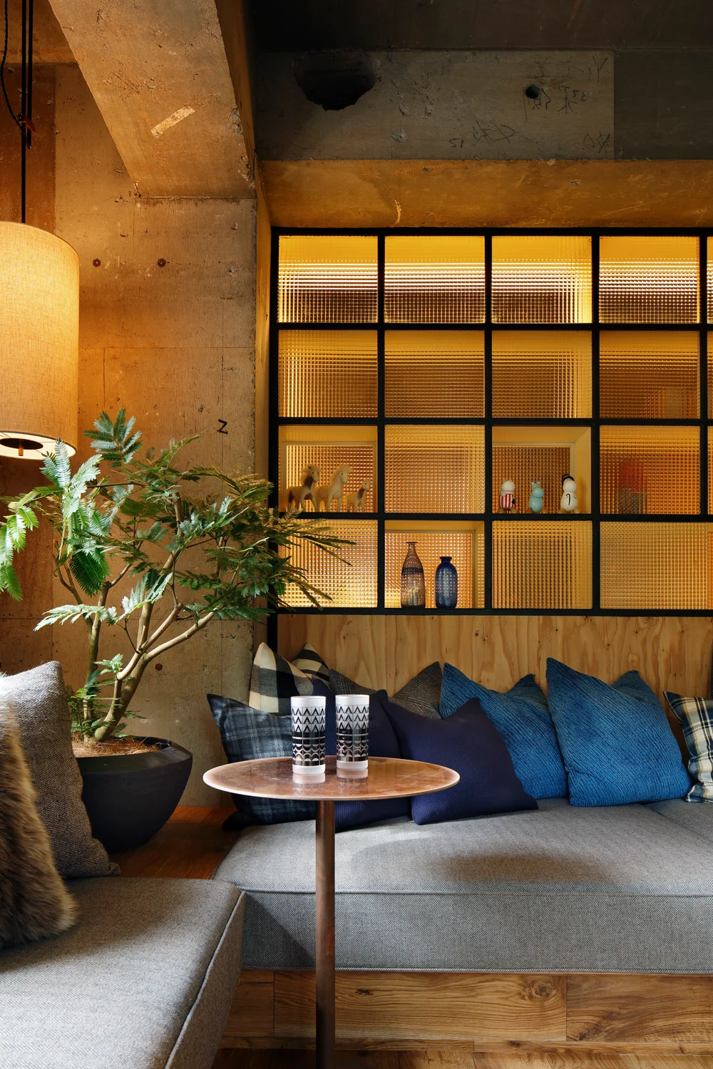 A built-in wooden sofa with grey and blue pillows sits against a raw concrete wall and a large backlit amber glass grid partition.