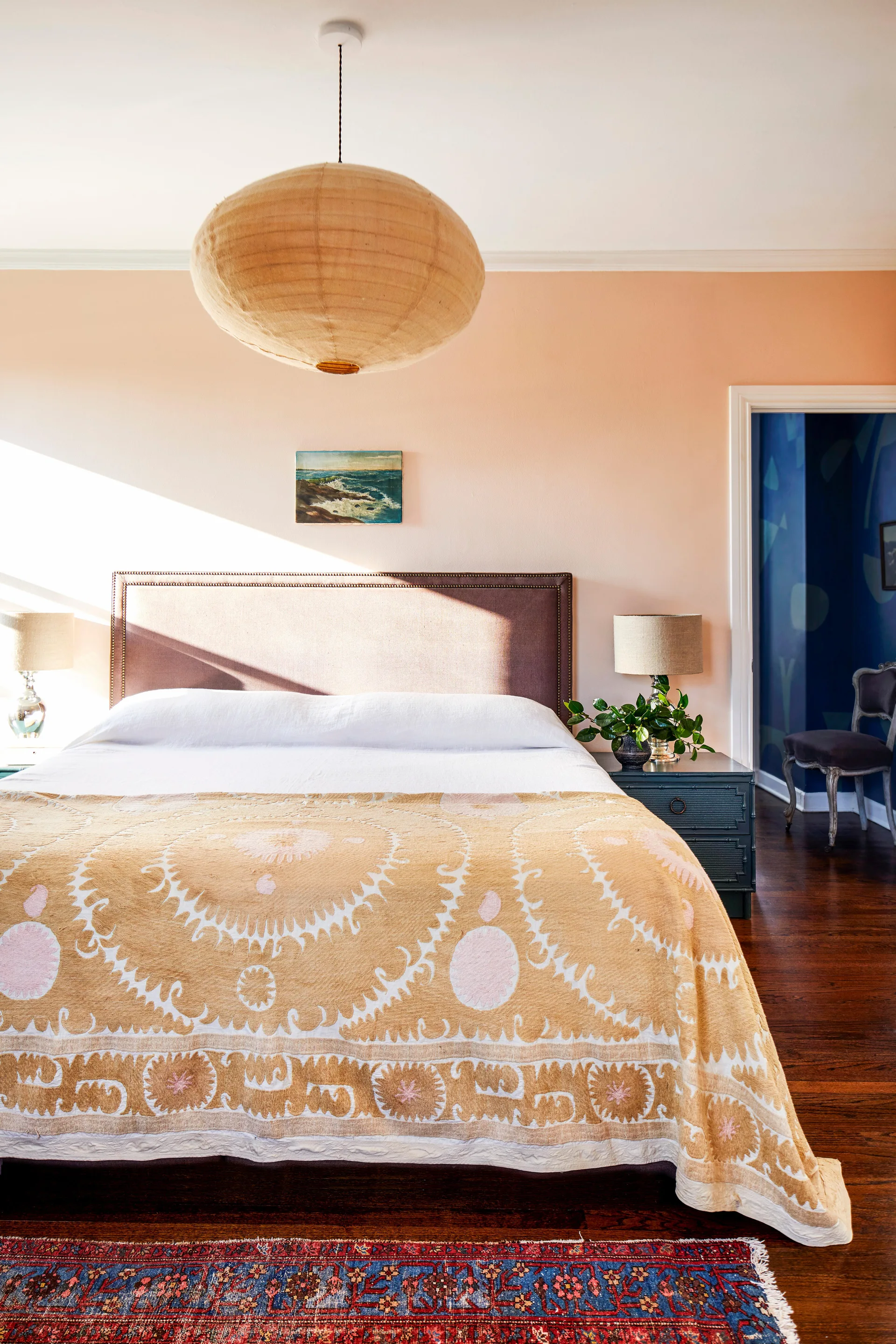 A bedroom with peach walls features a bed with a patterned yellow coverlet, a large paper pendant light, and a view into a vibrant blue room.