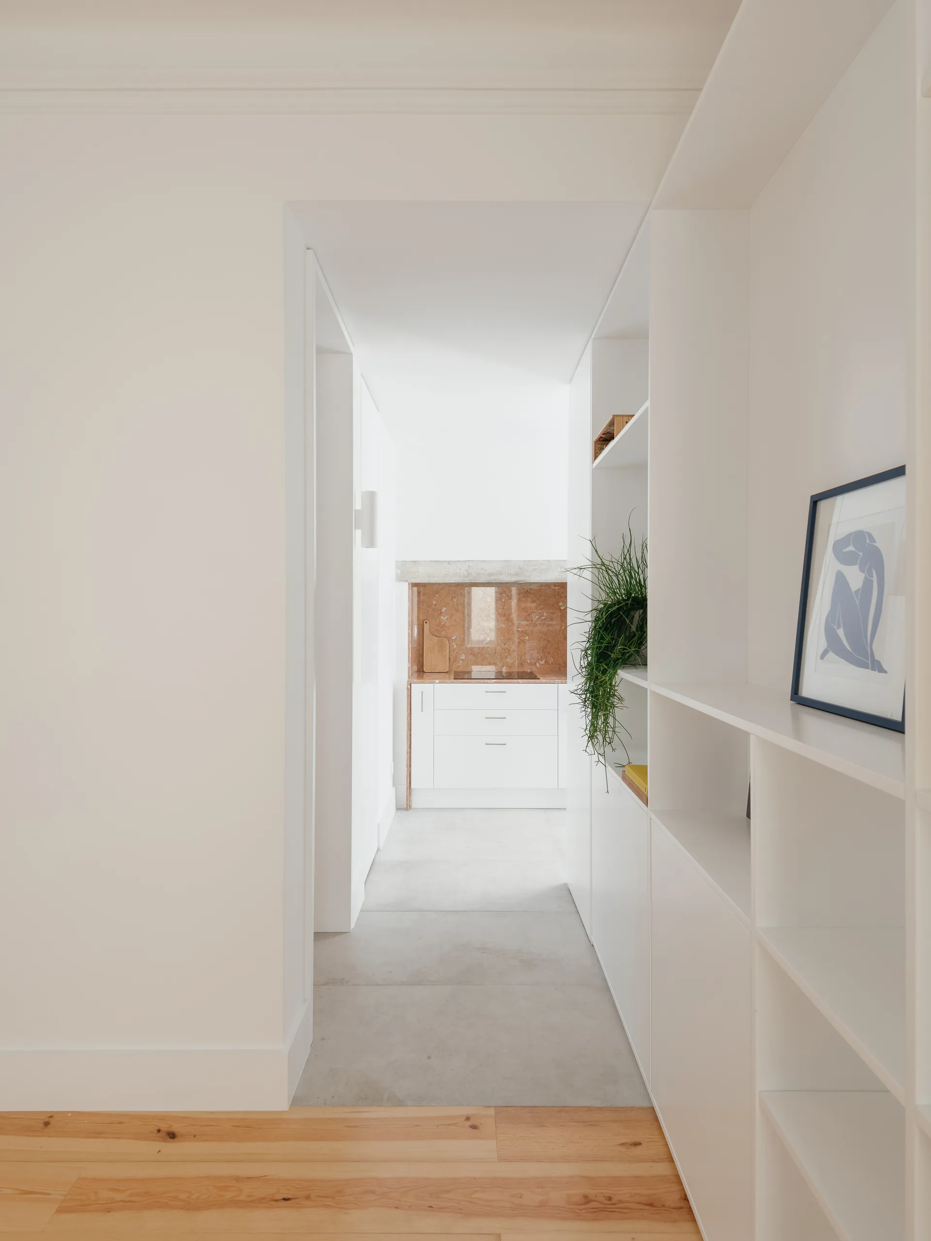 A perspective view of a white hallway with built-in shelving and a transition from wood to gray tile flooring, leading to a kitchen.
