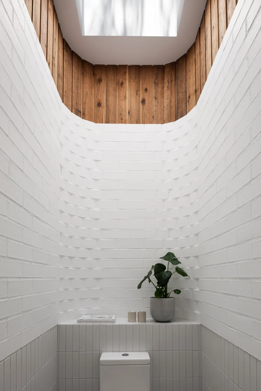A minimalist white bathroom features a curved brick wall, a skylight lined with vertical wood panels, and a small potted plant on a tiled ledge.