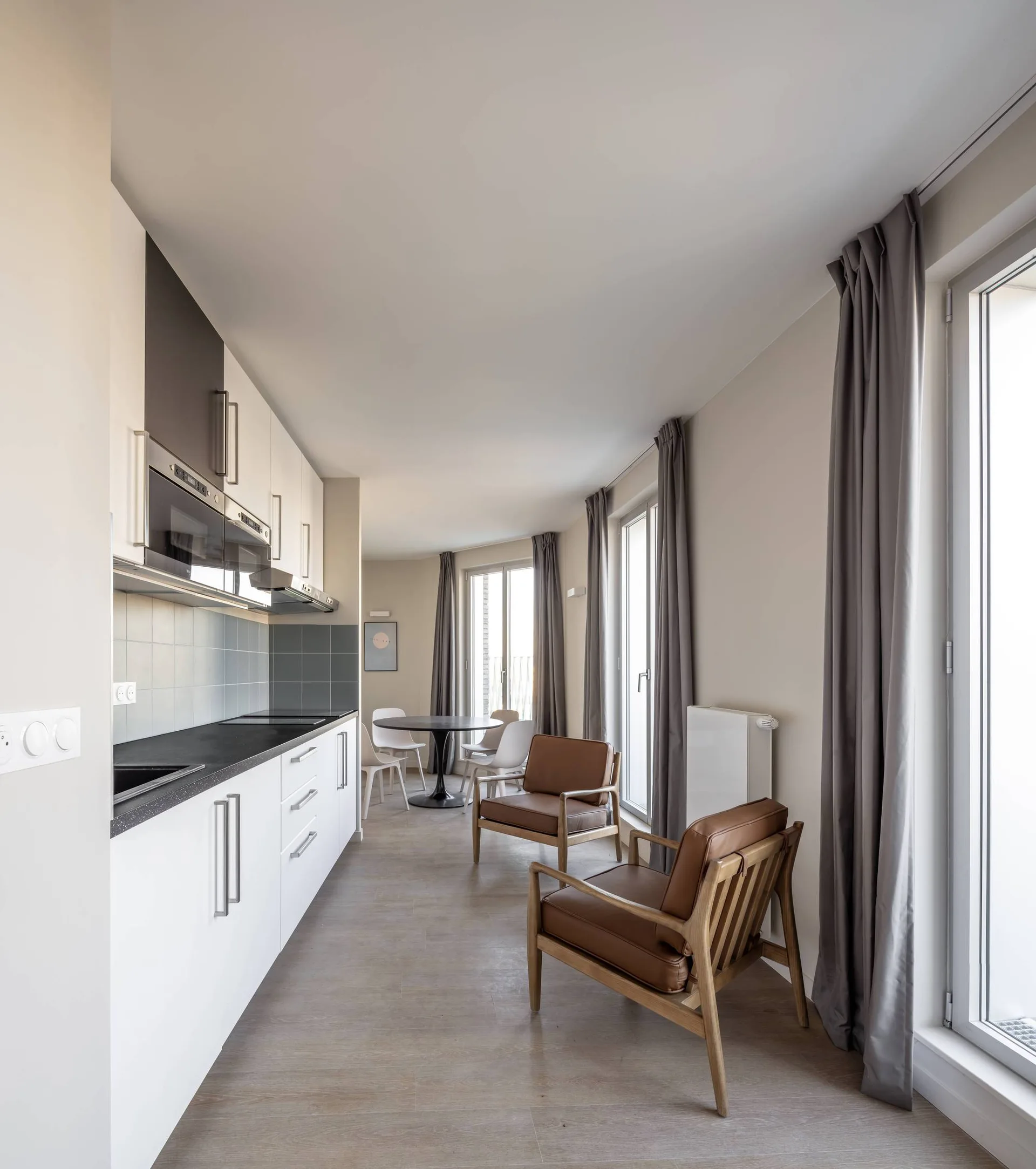 A modern galley kitchen with white cabinets and a dark countertop faces two brown leather armchairs in an open-plan apartment layout.