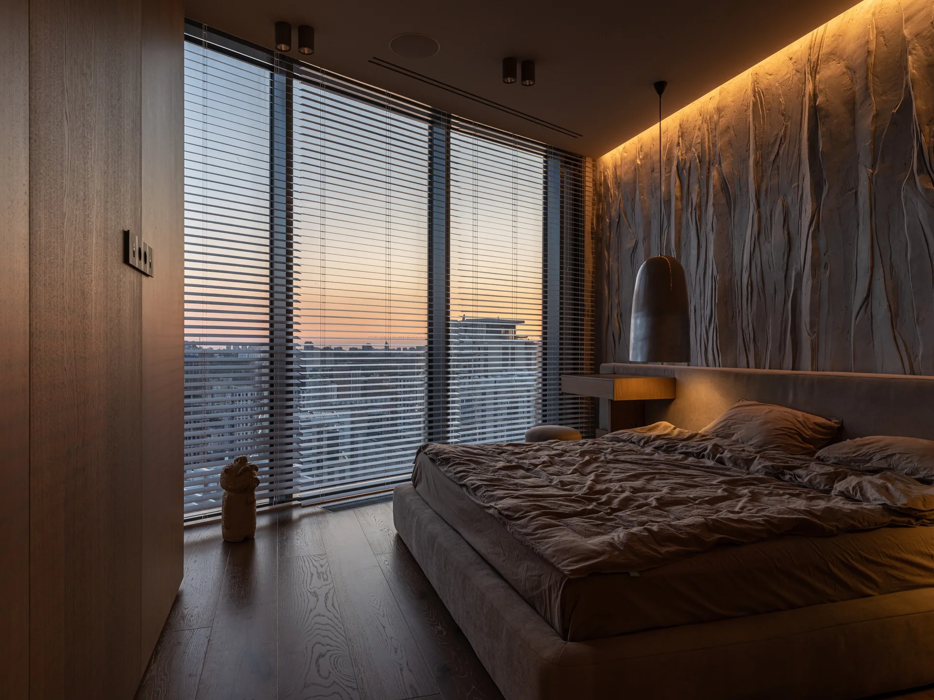 A minimalist bedroom features a large textured accent wall, a low platform bed, a metallic pendant light, and floor-to-ceiling windows with blinds.