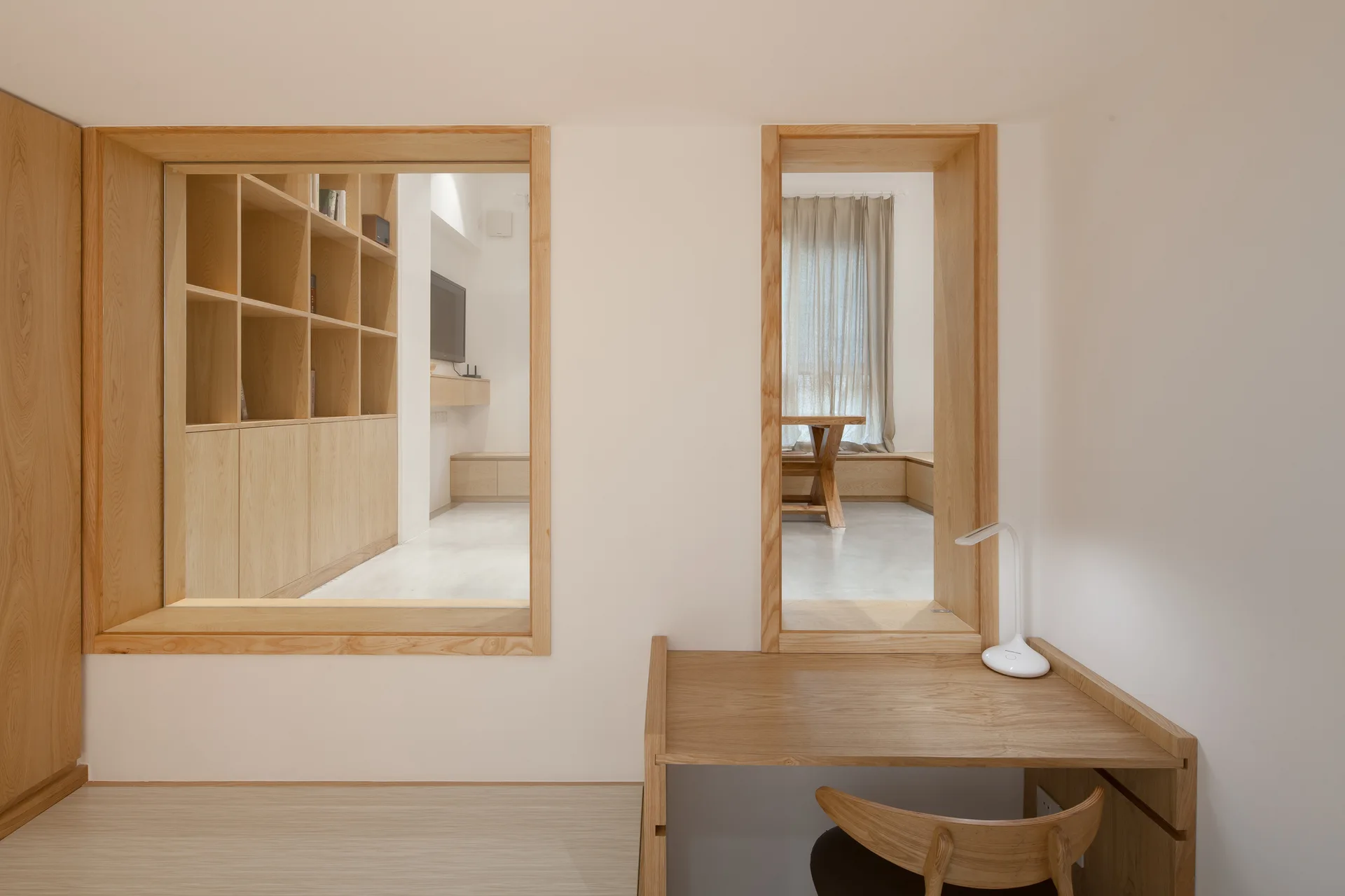 A light wood desk and curved chair sit before a white wall featuring two large, wood-framed square cutouts showing adjacent minimalist rooms.