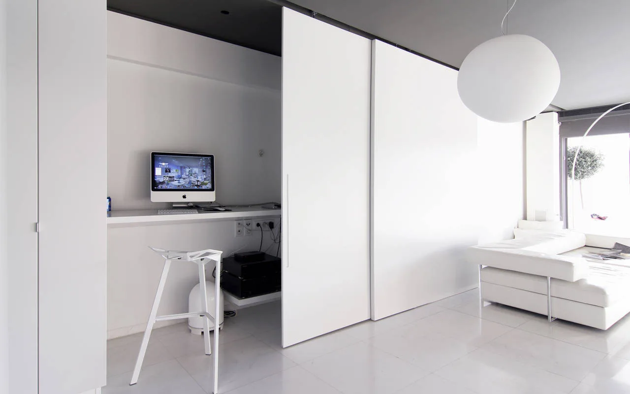 A modern interior features a built-in computer desk and stool partially concealed by large white sliding panels next to a white sectional sofa.