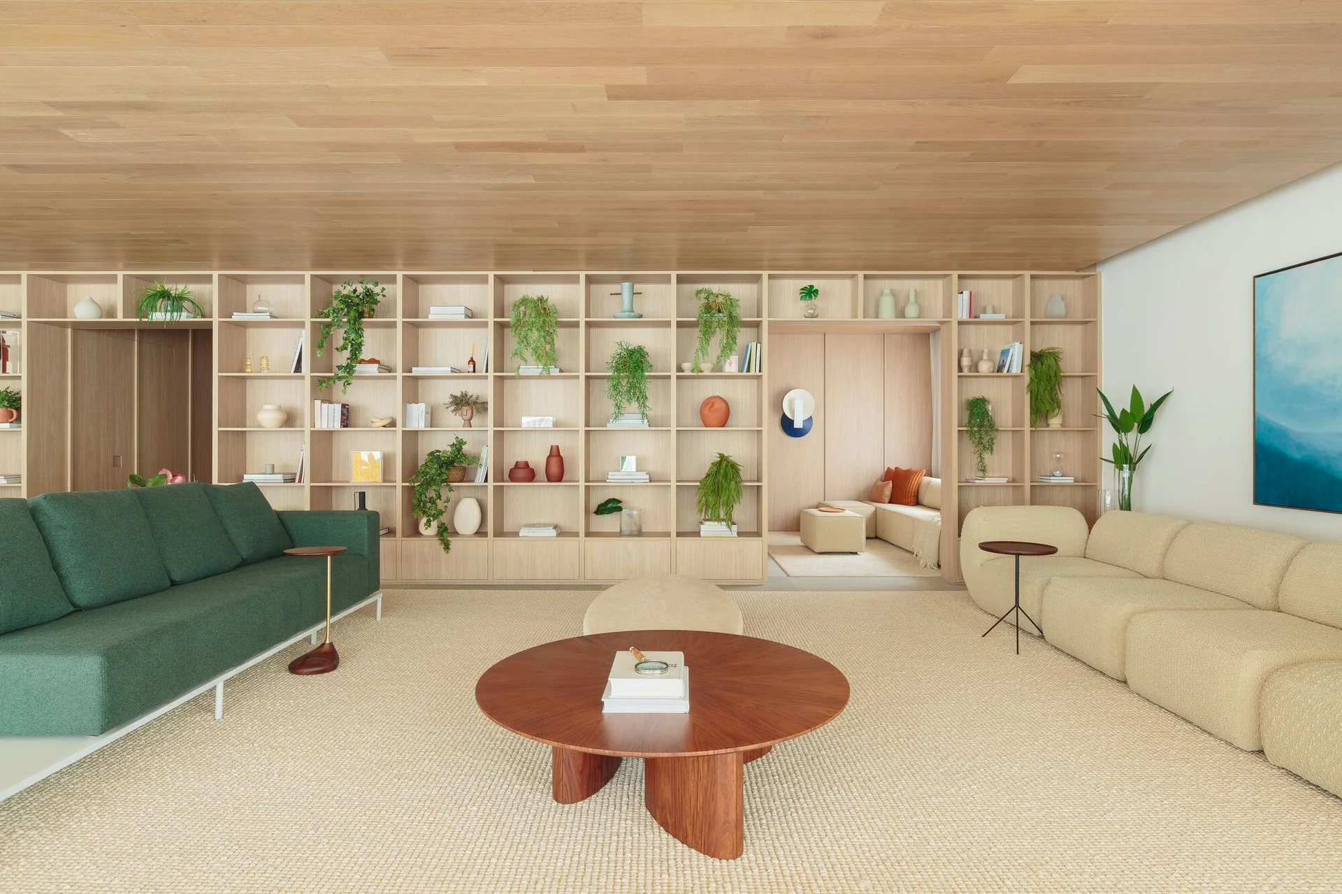 A spacious living room features green and beige sofas, a round wooden coffee table, and an expansive light wood shelving unit beneath a wood-paneled ceiling.