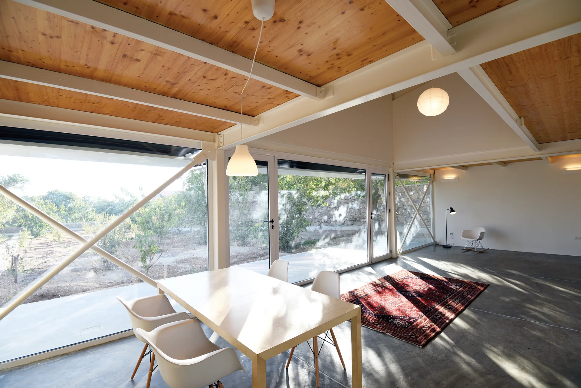 An open modern dining room featuring floor-to-ceiling windows with steel cross bracing, a light wood ceiling, and a polished concrete floor.