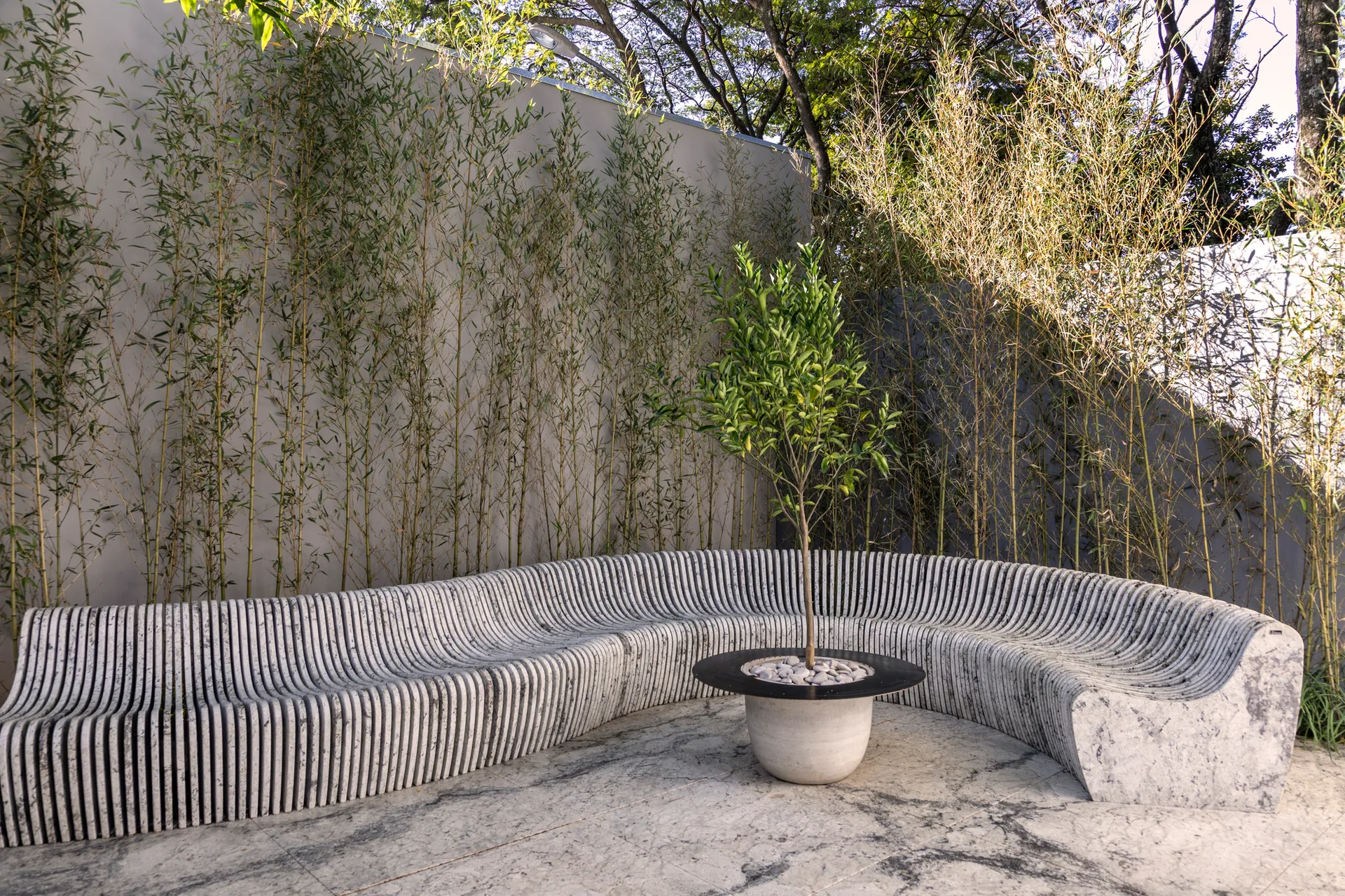 A large, curved bench made of slatted gray concrete sits on a stone tiled patio in front of a tall bamboo privacy screen.